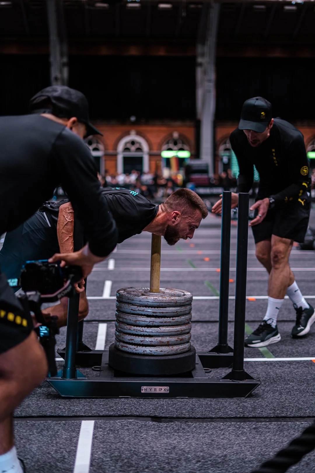 A man participating in a strongman event, pushing a large weight on a wooden sled while being assisted by two spotters inside a gym or event hall.