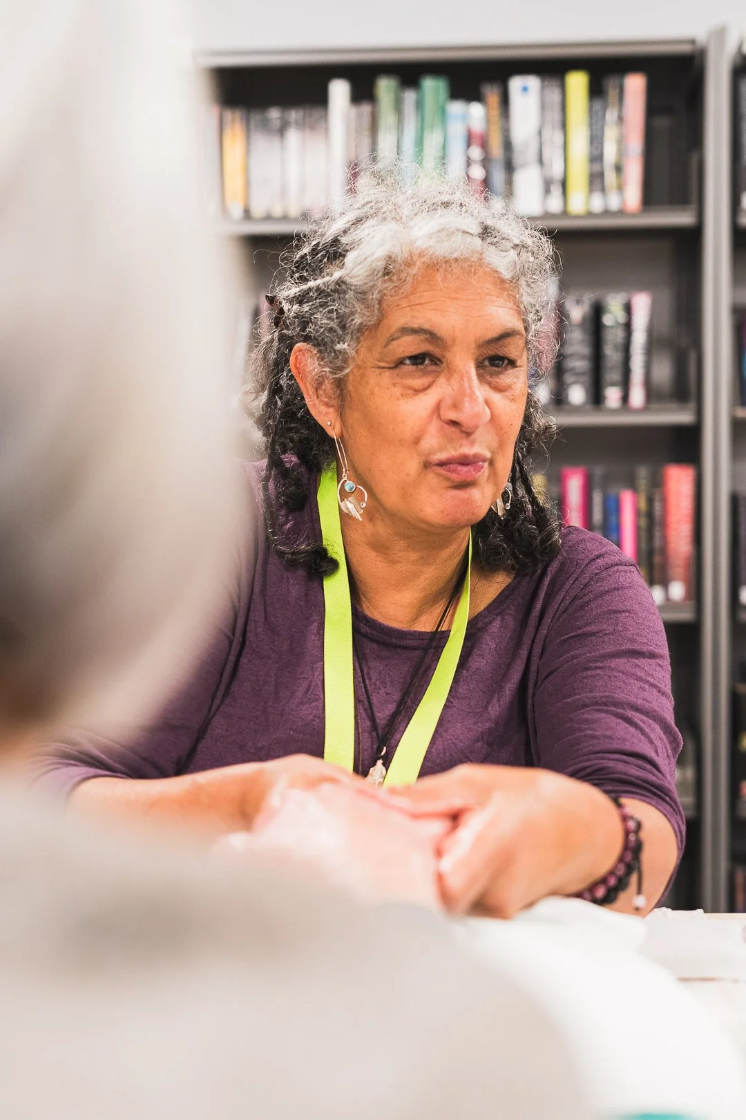 A woman with curly gray hair and earrings, wearing a purple shirt and a bright green lanyard, seated at a table with a background of bookshelves filled with books.