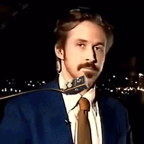 Man in suit with a mustache and beard, sitting in a dark room with city lights visible in the background, and a microphone attached to his neck.