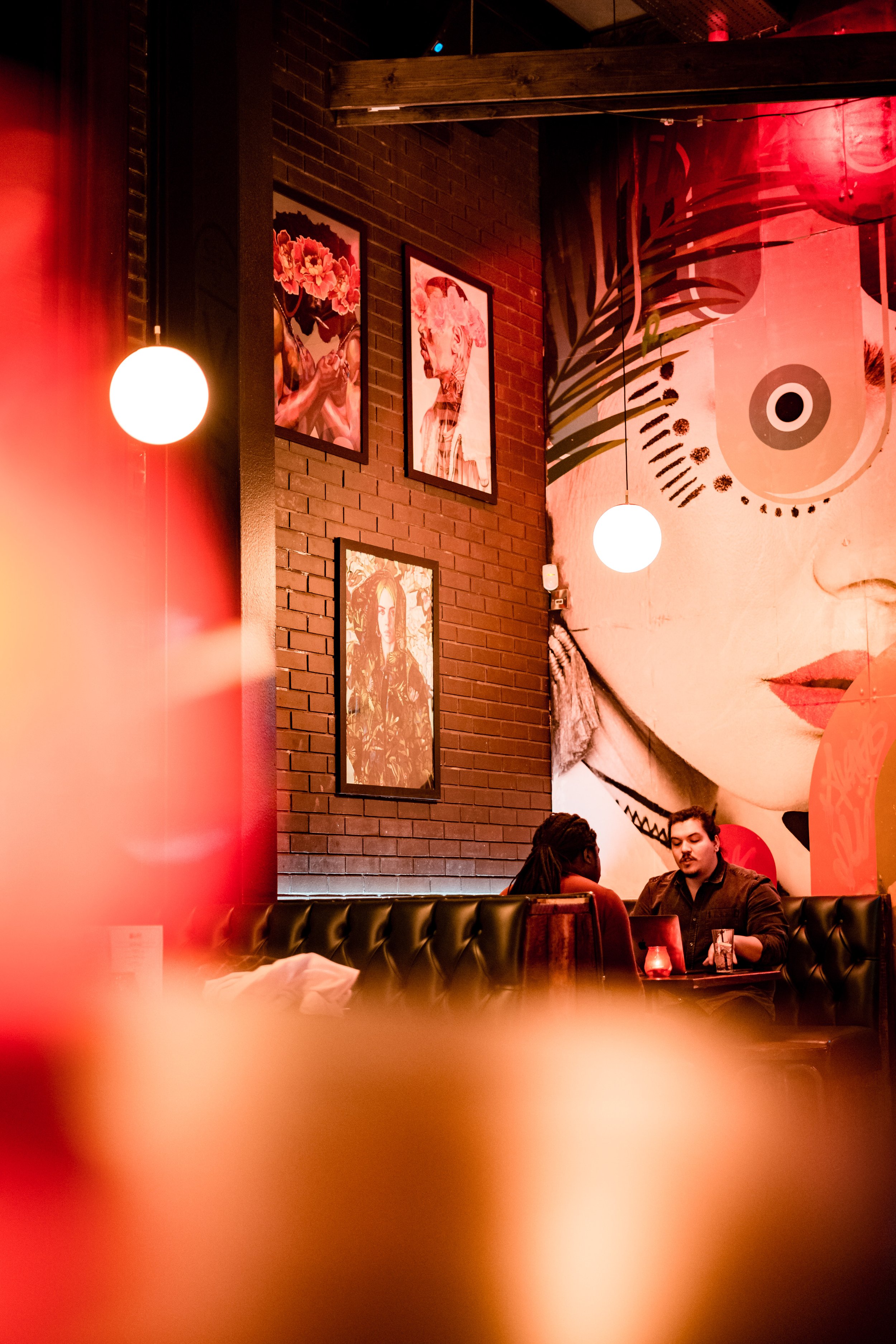 A cozy restaurant interior with warm lighting, featuring artwork on brick walls and two people sitting at a table engaged in conversation.