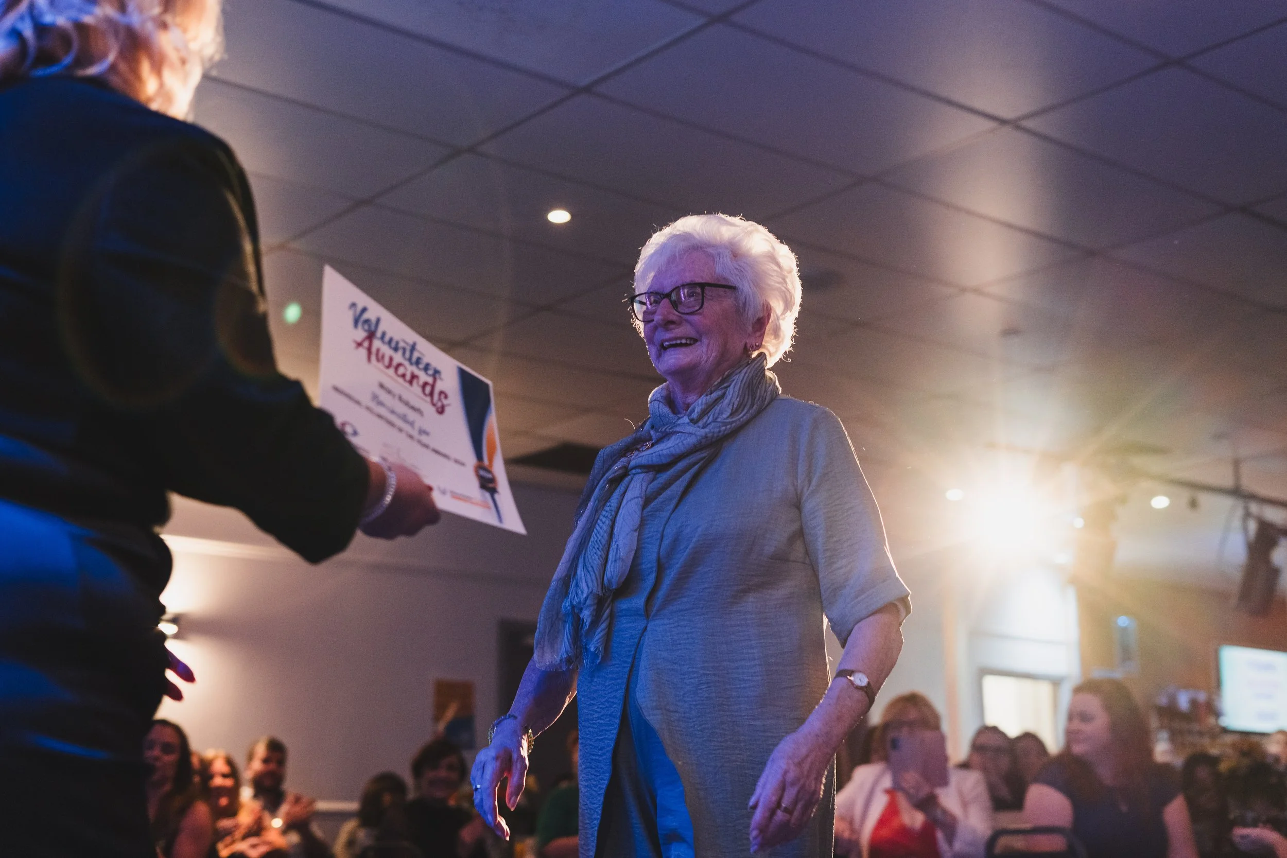 An elderly woman with white hair, glasses, and a gray scarf is smiling and receiving a certificate or award at an indoor event, with several people seated in the background.