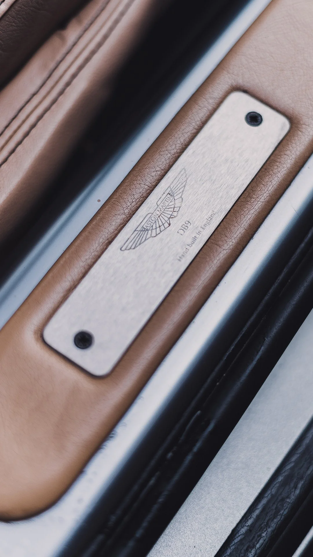 Close-up of a metal plaque on a luxury car door frame with the Aston Martin logo, model D9, and the inscription 'hand built in England'.