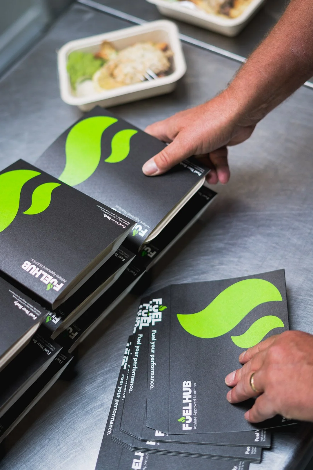 Hands handling FUELHUB flyers with green leaf logo, on a table near food containers.