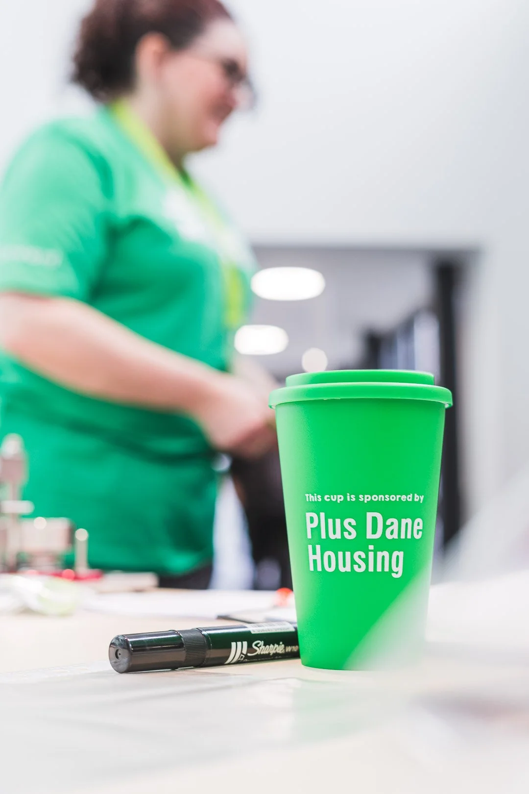Close-up of a green paper cup with the text 'This cup is sponsored by Plus Dane Housing' placed on a table next to a black Sharpie marker. In the blurred background, a woman wearing glasses and a green shirt is standing.