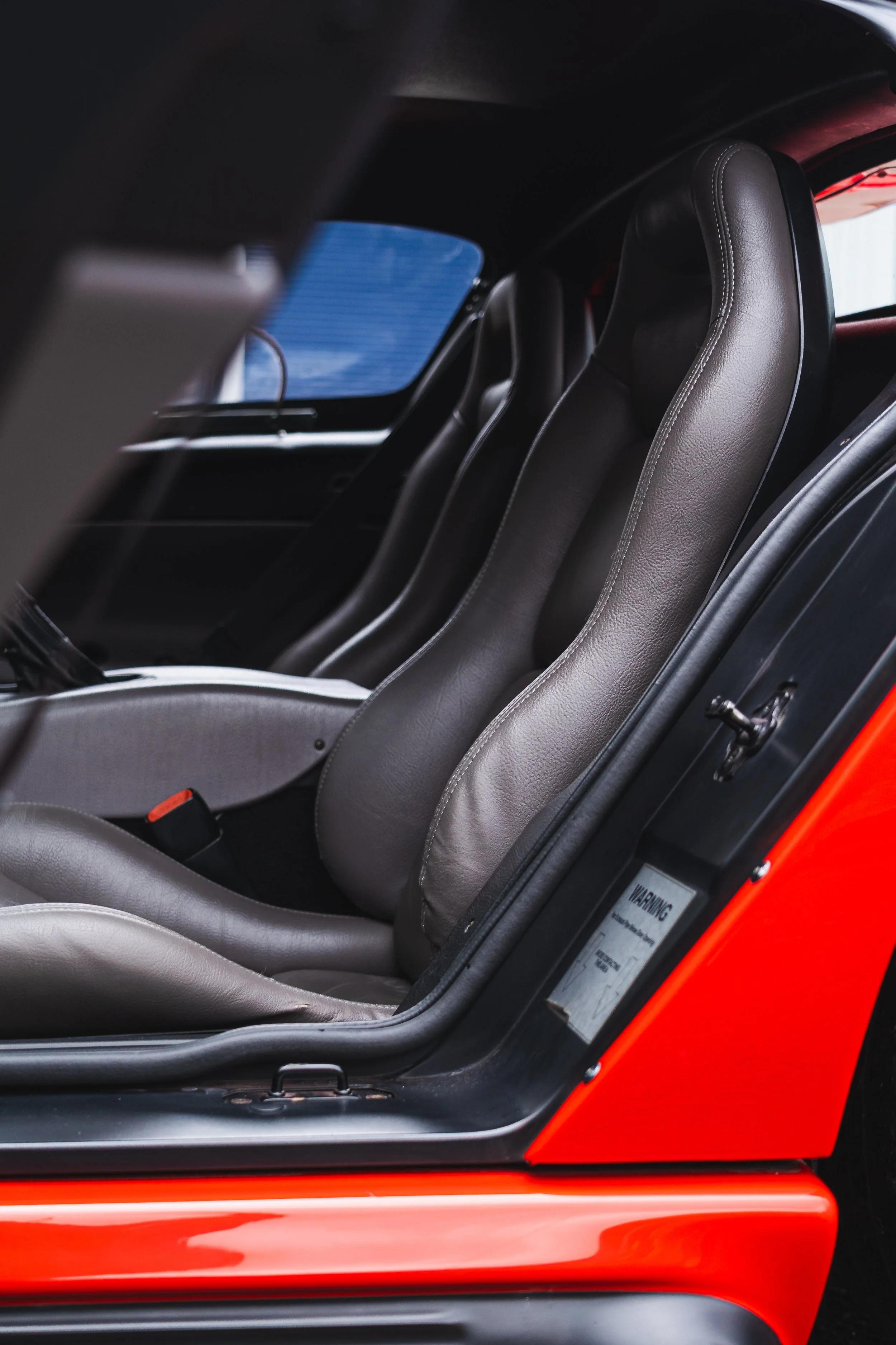 The interior of a race car shows gray leather racing seats in a side view, with a red exterior visible on the right edge.