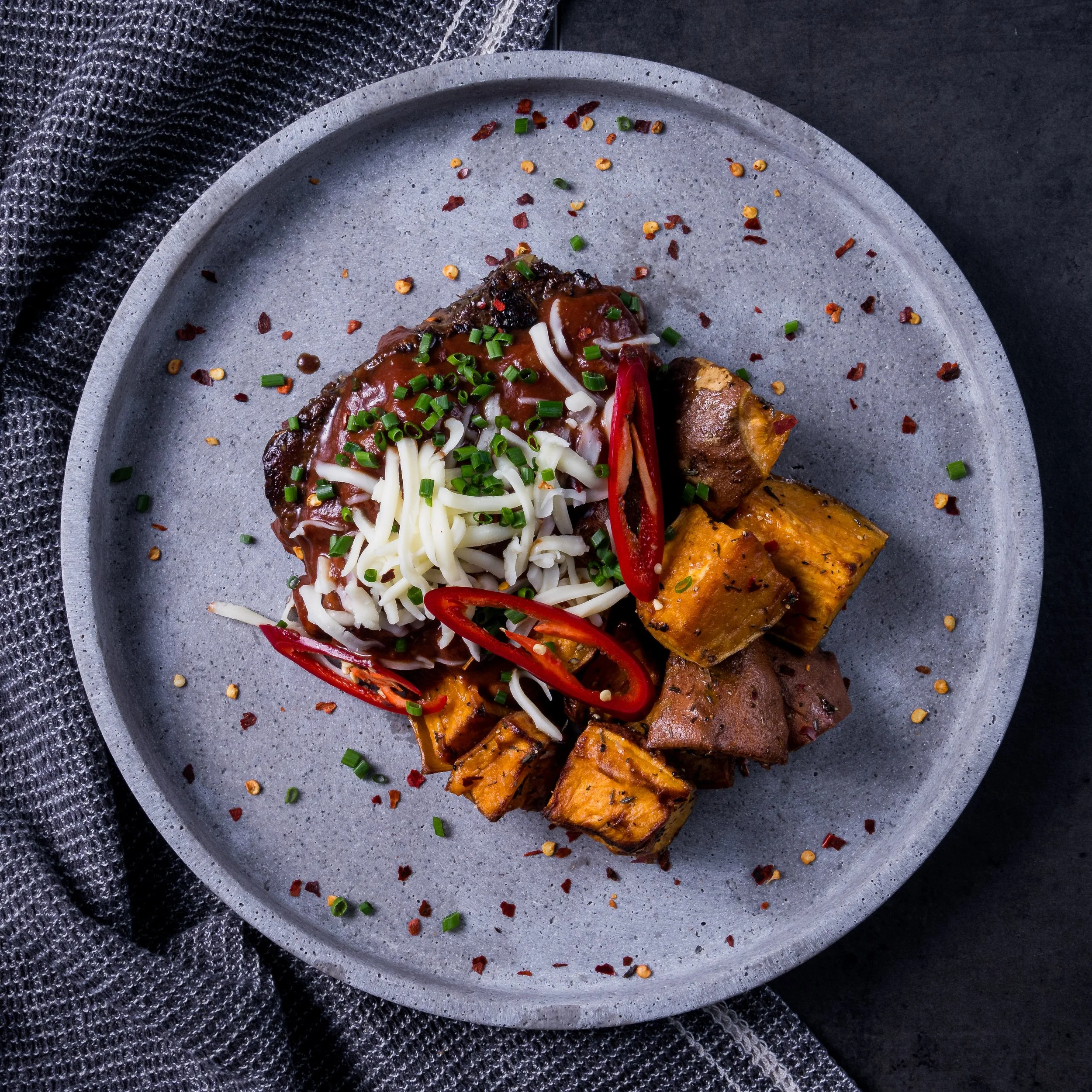 A plate of sweet potato cubes, beef chunks, and a meatball with gravy, topped with shredded cheese, chopped chives, and sliced red chili peppers, garnished with red pepper flakes and sesame seeds.