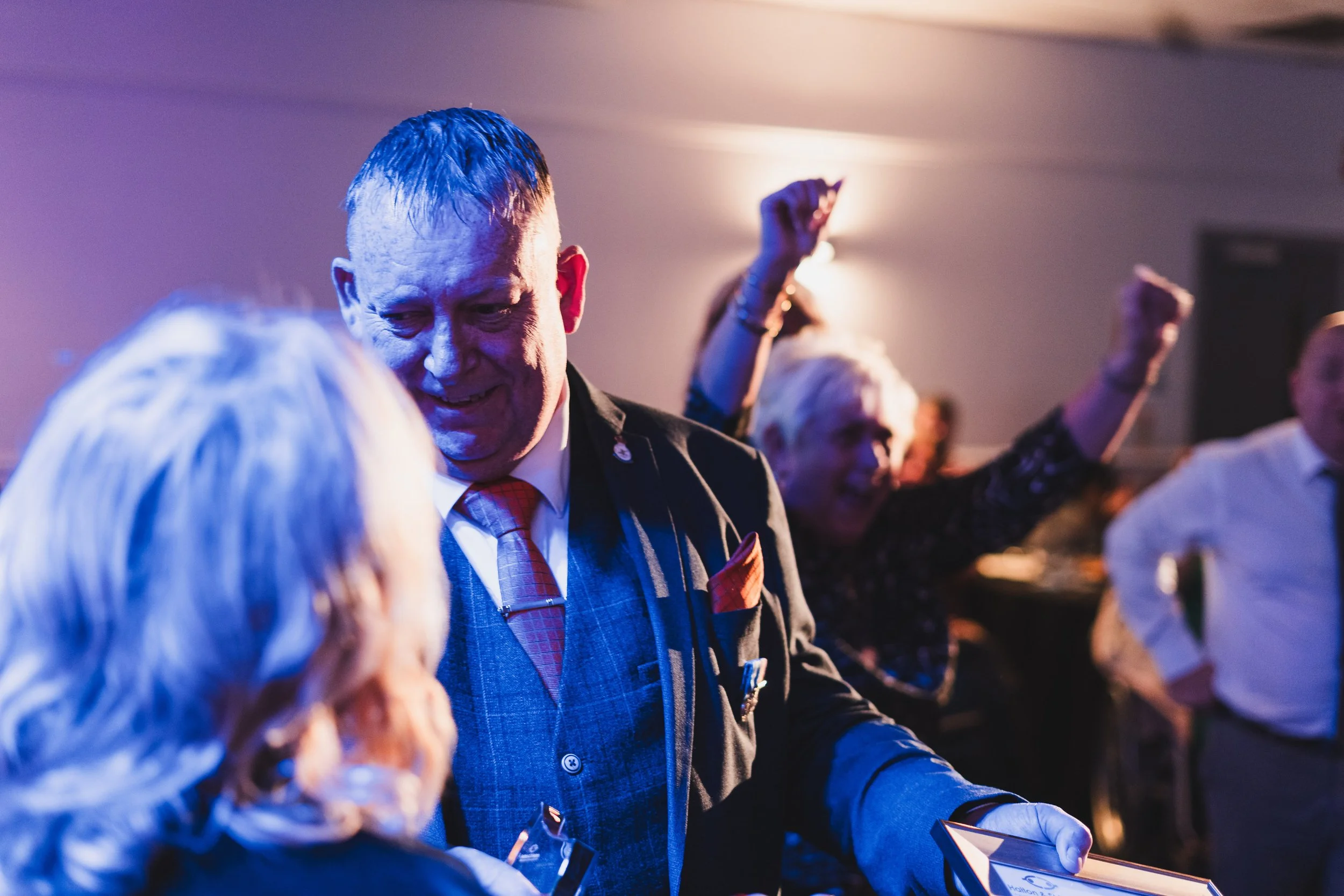 People dancing and smiling at a party, with one man in a suit and a woman with white hair in the foreground.