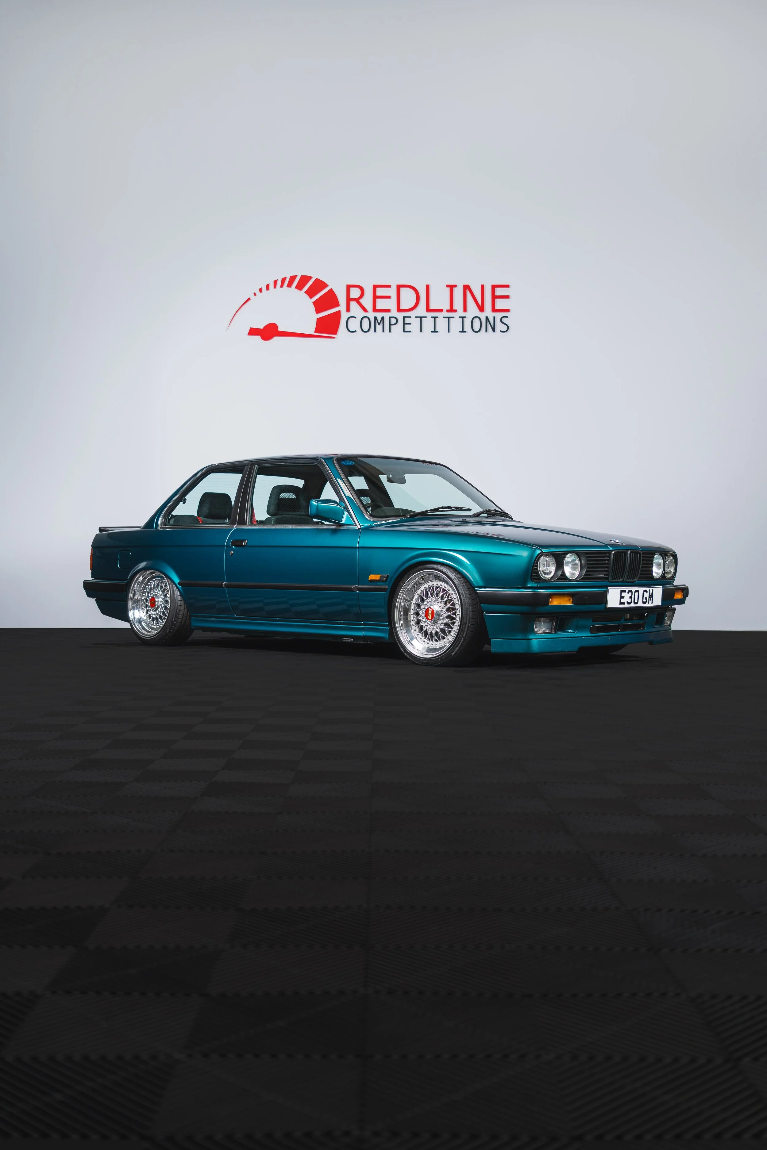 A vintage teal BMW E30 coupe with custom wheels parked indoors in front of a white wall with the 'Redline Competitions' logo.