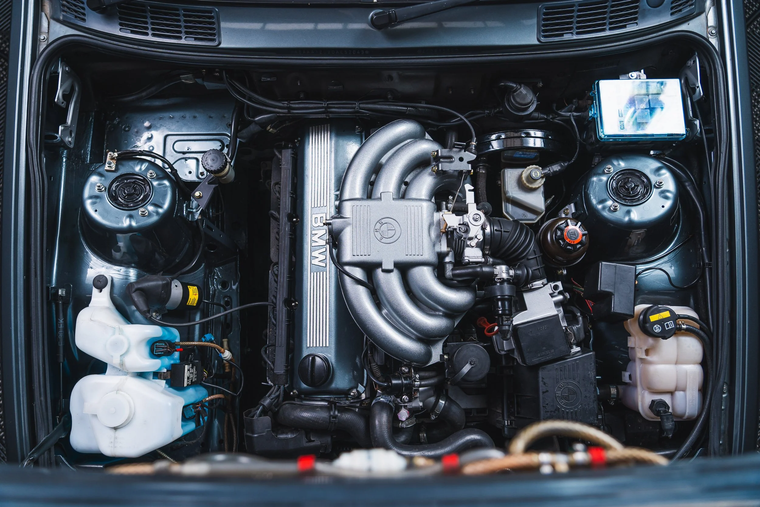 Top view of a car engine bay featuring a BMW engine, various hoses, containers, and components.