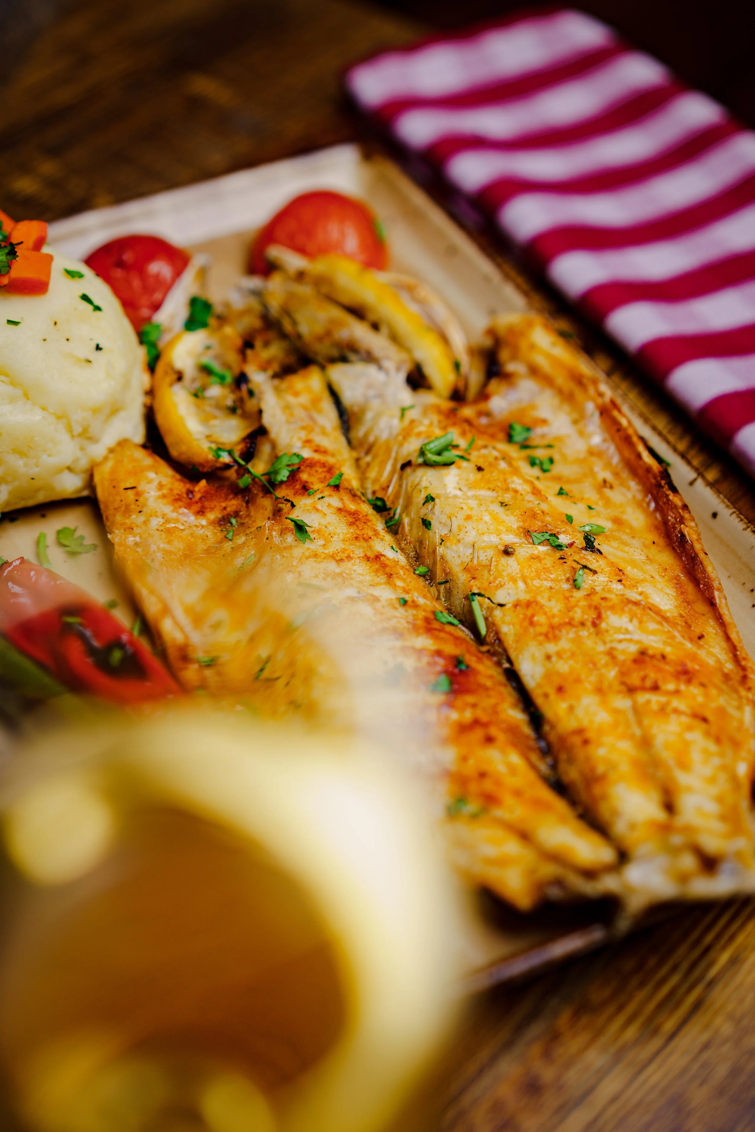 A plate with grilled fish fillets garnished with chopped herbs, mashed potatoes with vegetables, cherry tomatoes, and roasted vegetables on a wooden table, with a striped napkin nearby.