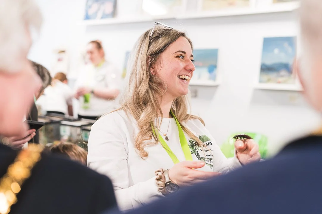 A woman with blonde hair smiling and talking to two people, at an indoor event with artwork on the walls in the background.