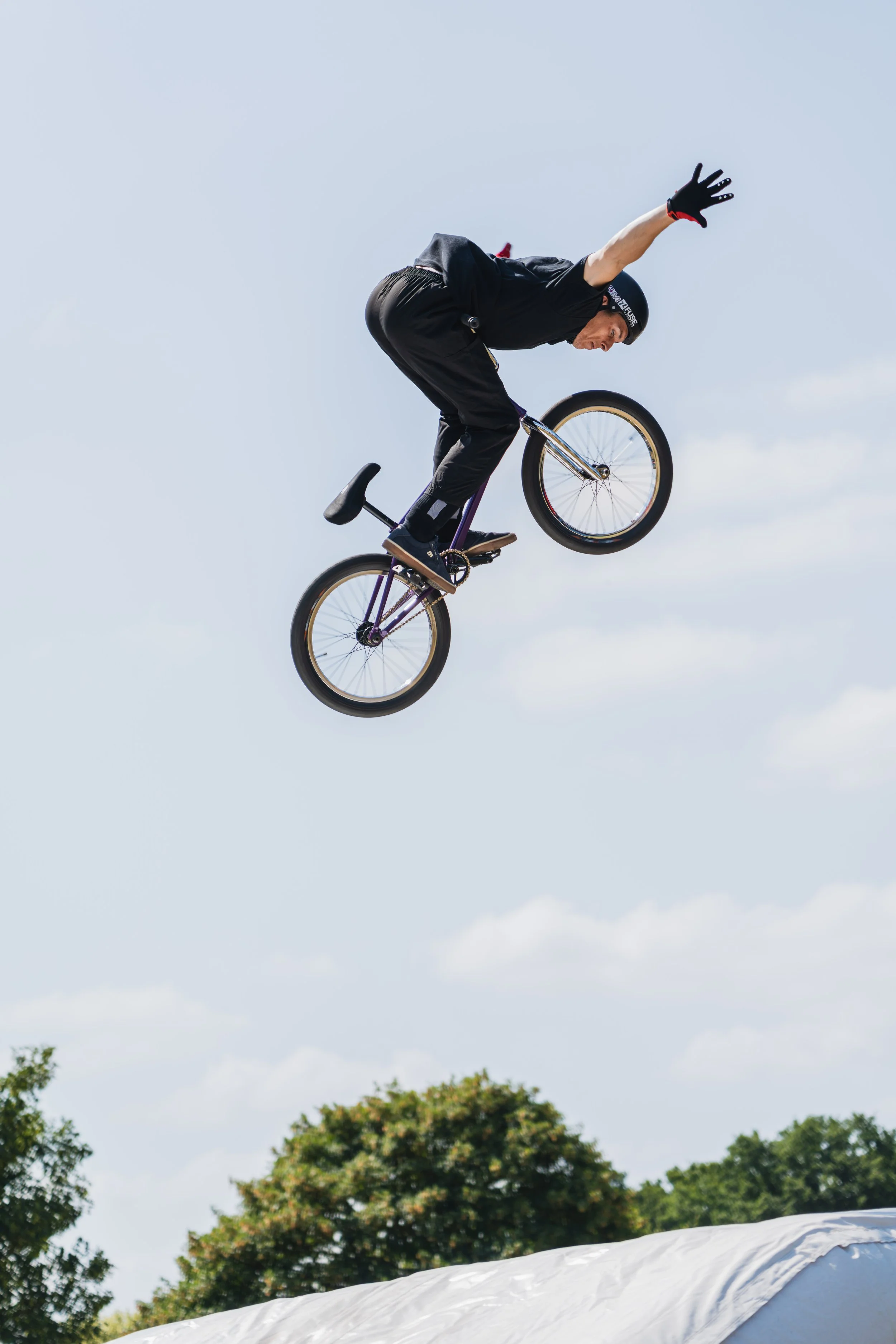 A person performing a jump on a BMX bike in mid-air against a cloudy sky. The person is wearing protective gear including a helmet and gloves, with trees visible in the background.