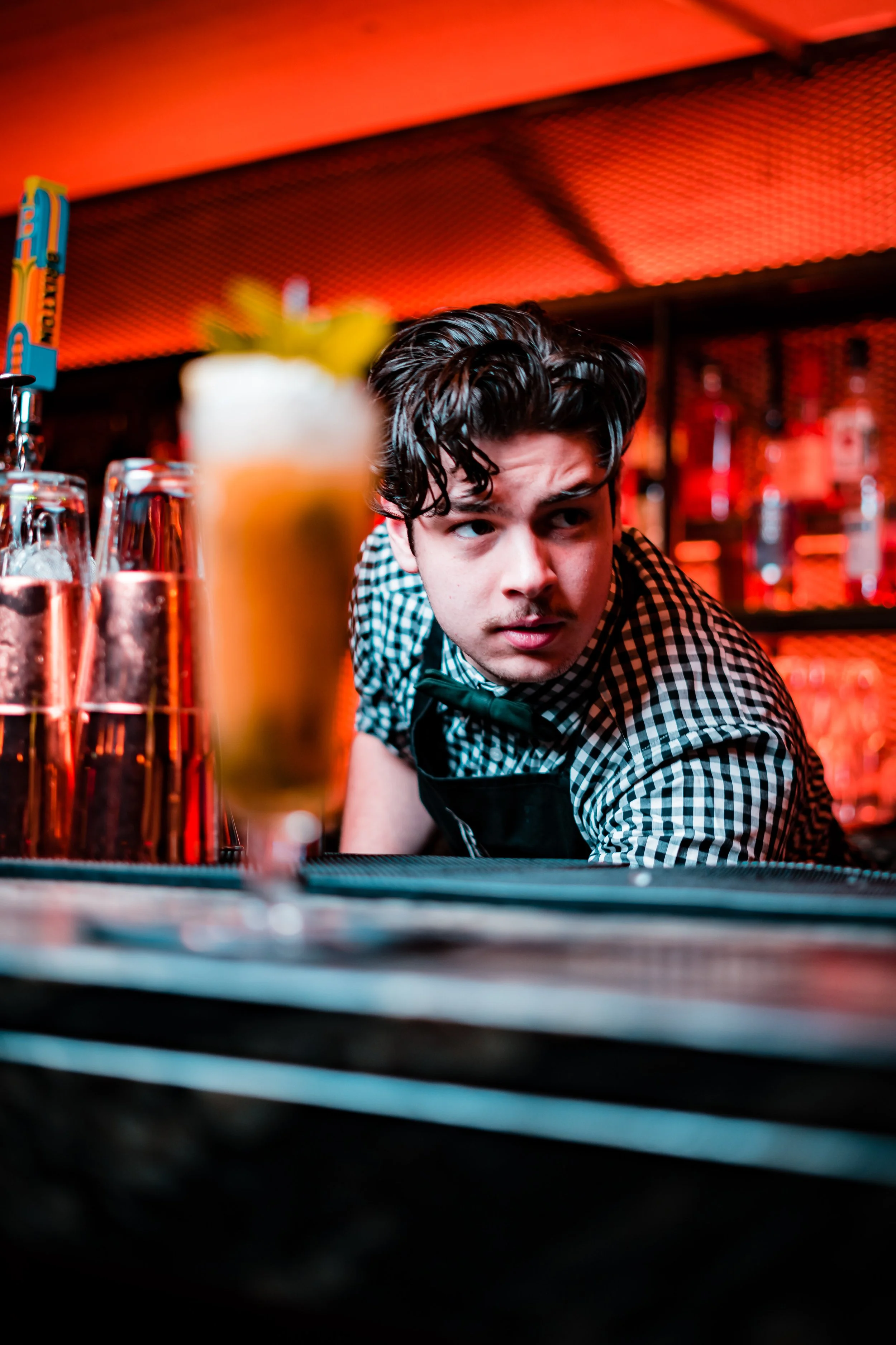 A bartender with curly dark hair wearing a black and white checkered shirt and a black apron behind a bar, looking to the side. There are glasses with beverages in the foreground, with a blurred background of bottles and bar equipment.