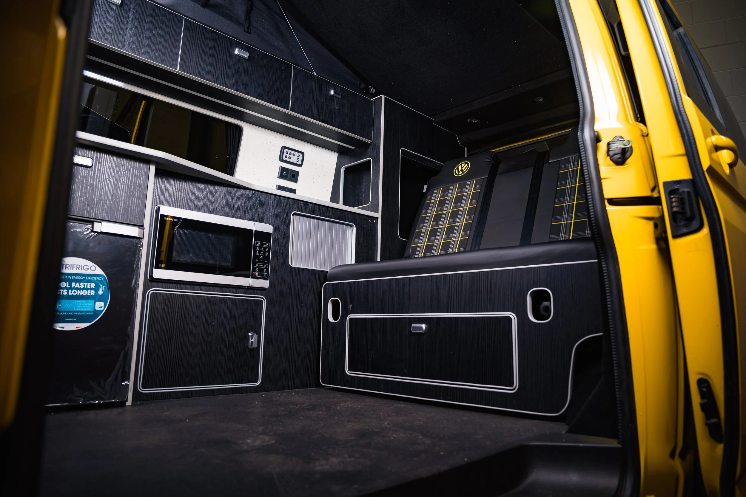 Interior of a van converted into a camper with black cabinetry, a small microwave, a seating area with plaid fabric, electrical outlets, and a fridge.