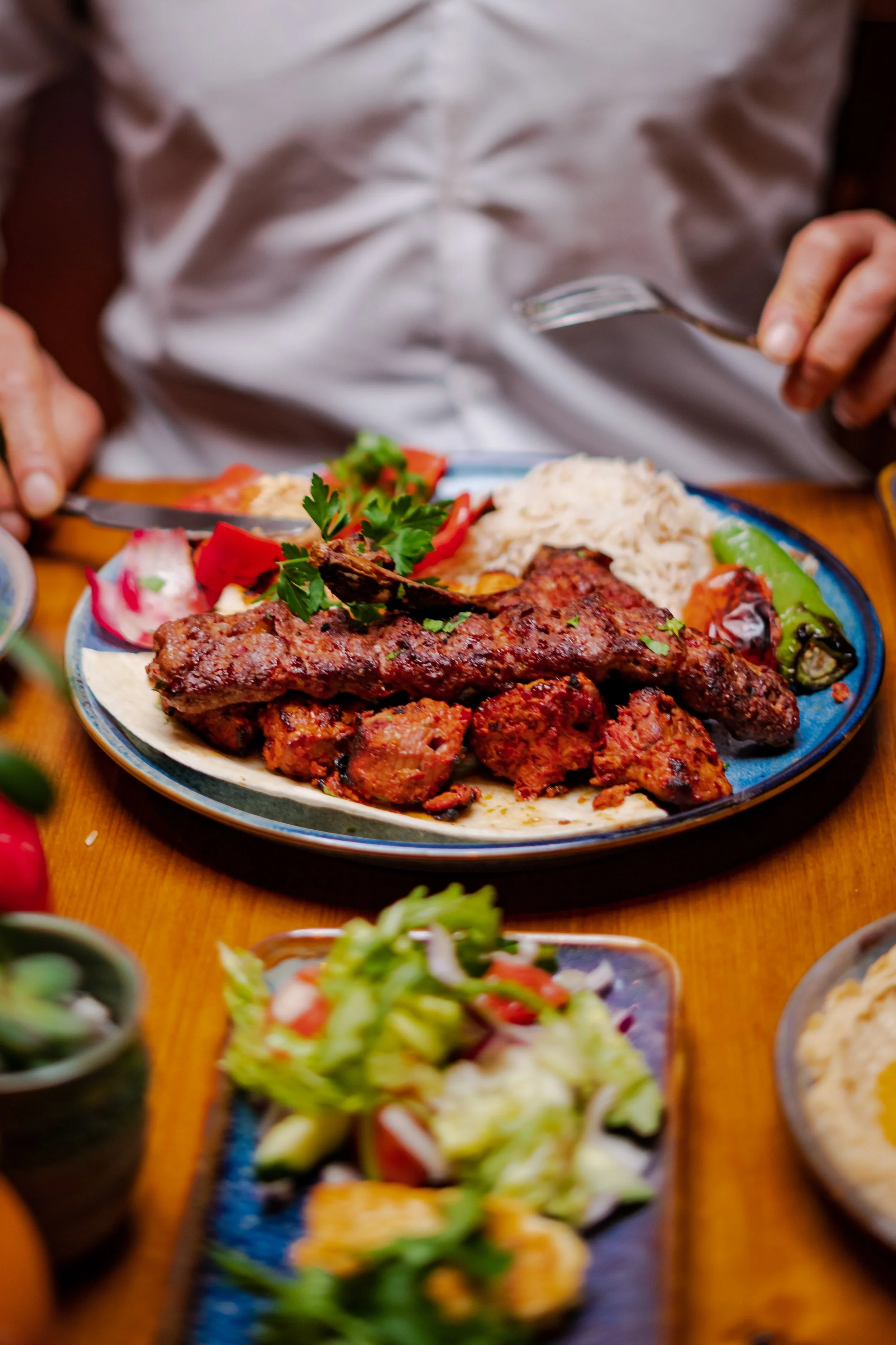 Plate of grilled meat, rice, and vegetables on a wooden table with a person holding a fork and knife.