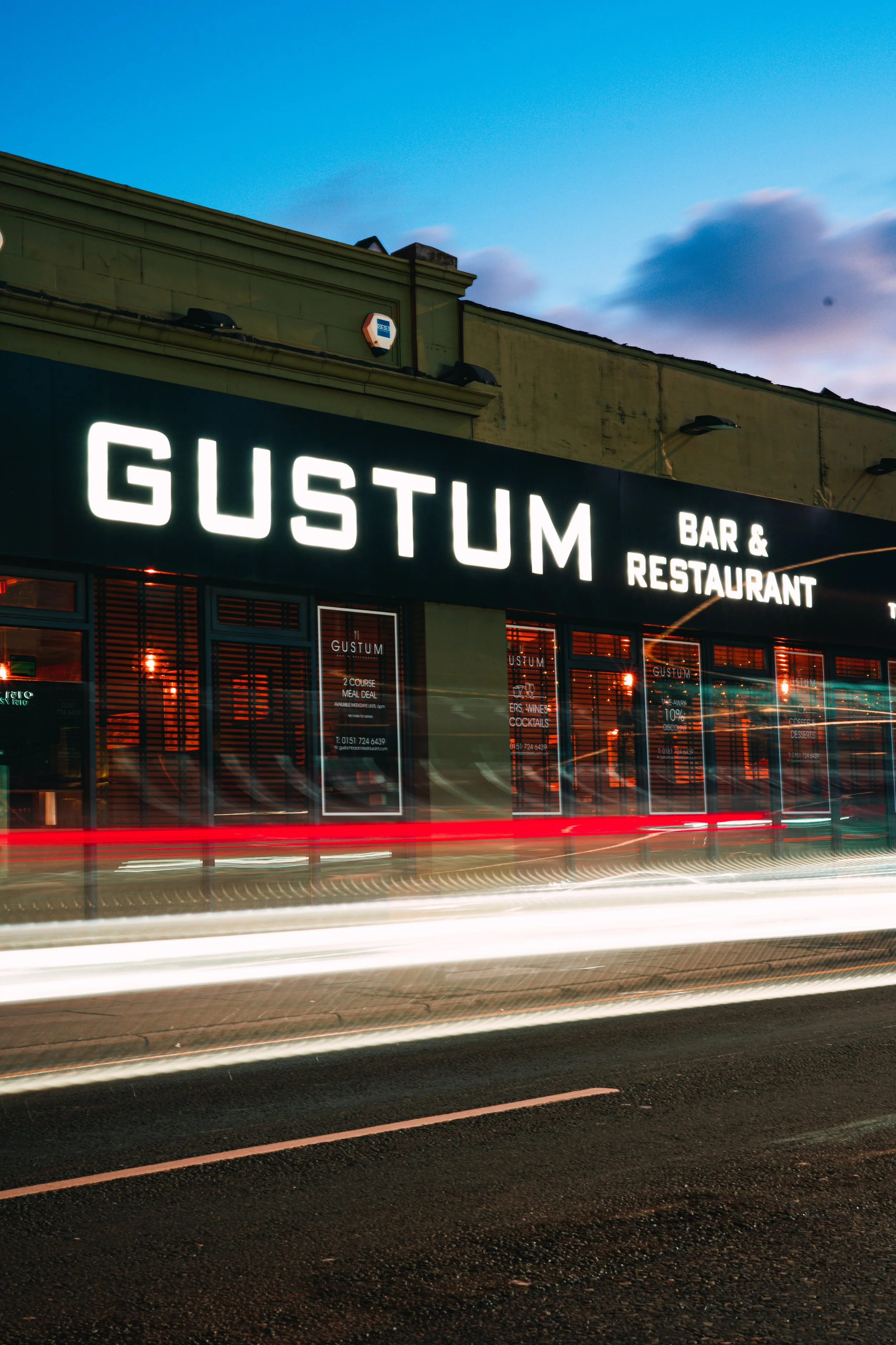 Nighttime photo of Gustum Bar & Restaurant storefront with illuminated signage, parked cars, and light trails from passing vehicles on the street.