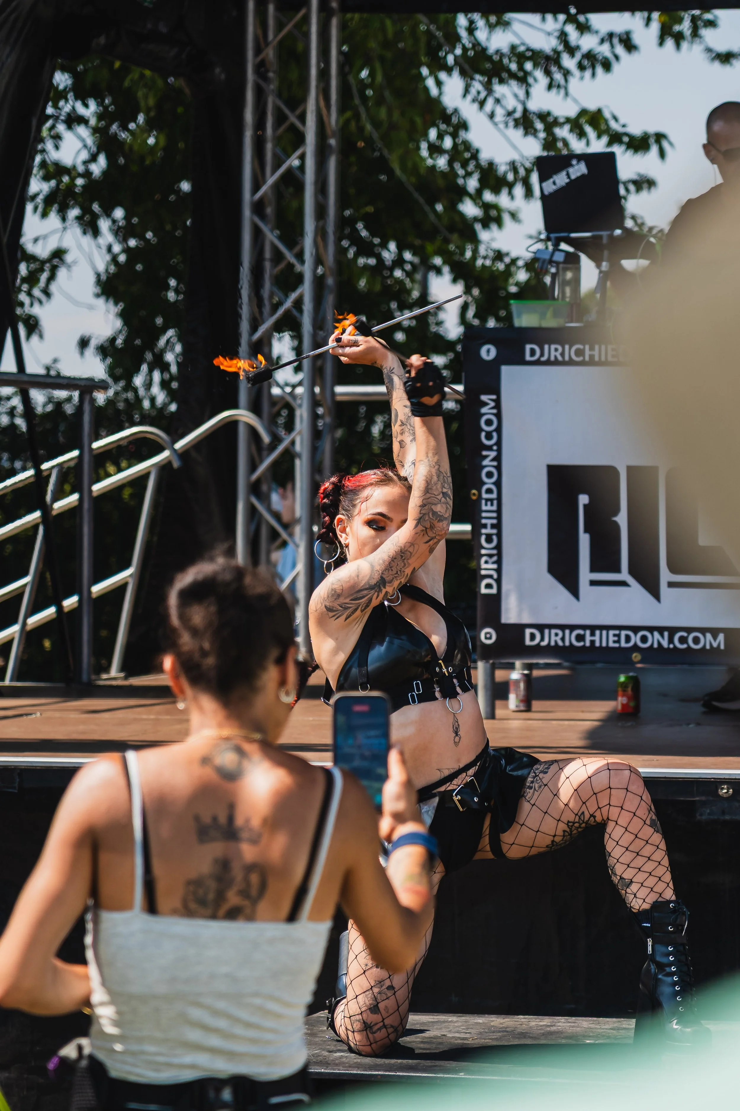 A performer with tattoos and fishnet stockings performing fire spinning on stage at an outdoor event, with a DJ and audience members watching.