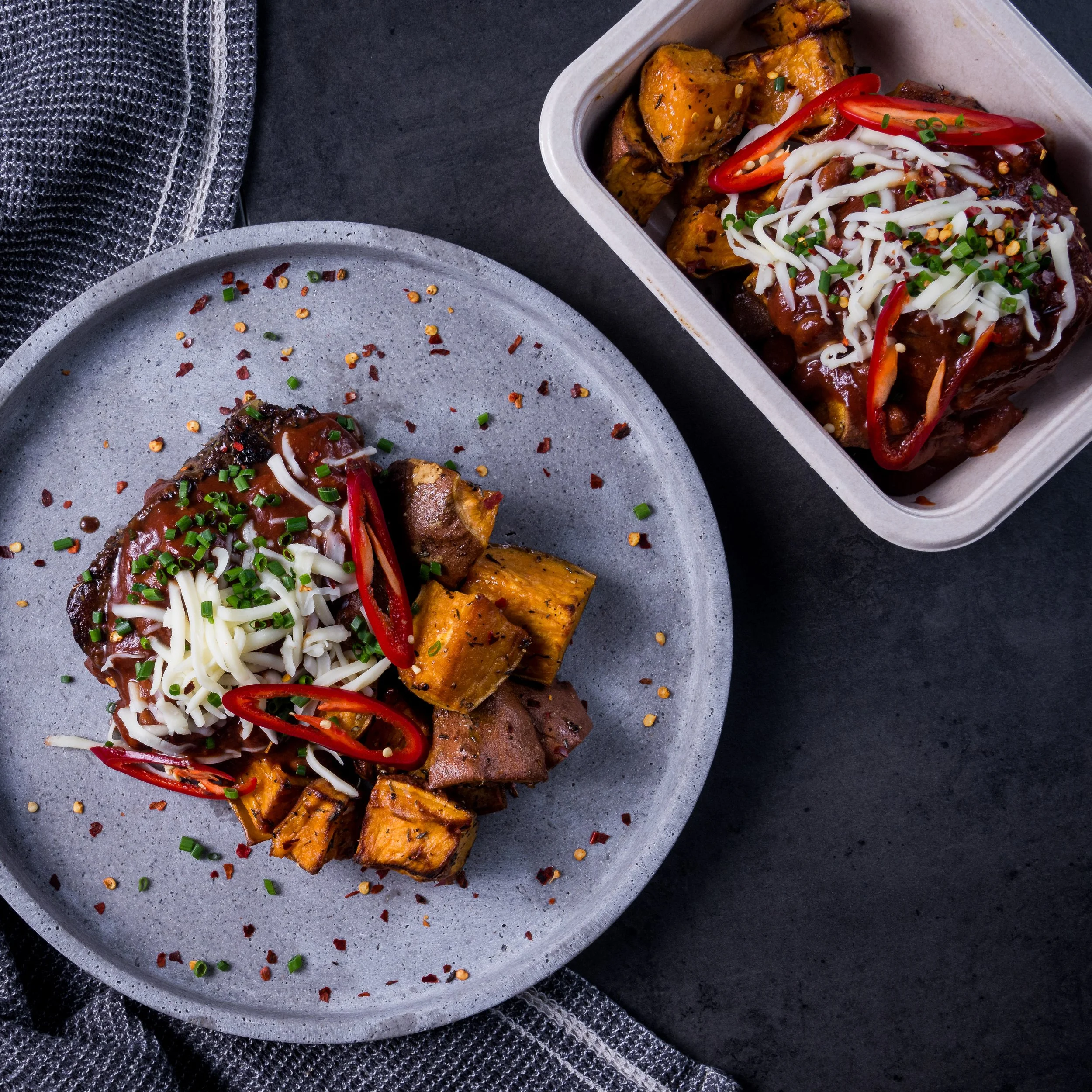 Plate and container of beef short ribs topped with shredded cheese, sliced red chili peppers, chopped chives, and red pepper flakes, served with roasted sweet potatoes on a dark surface.