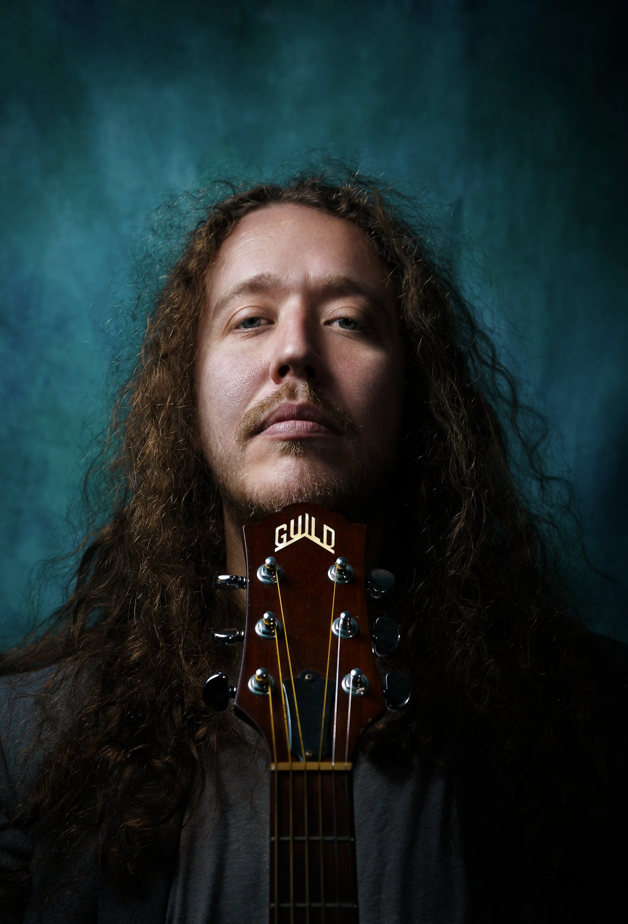 Portrait of a man with long curly hair and a mustache, holding a guitar headstock with the word 'GUILD' visible, against a dark teal background.