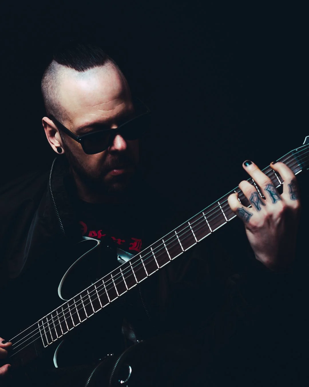 A man with a shaved head, wearing sunglasses and black clothing, playing an electric guitar against a dark background.
