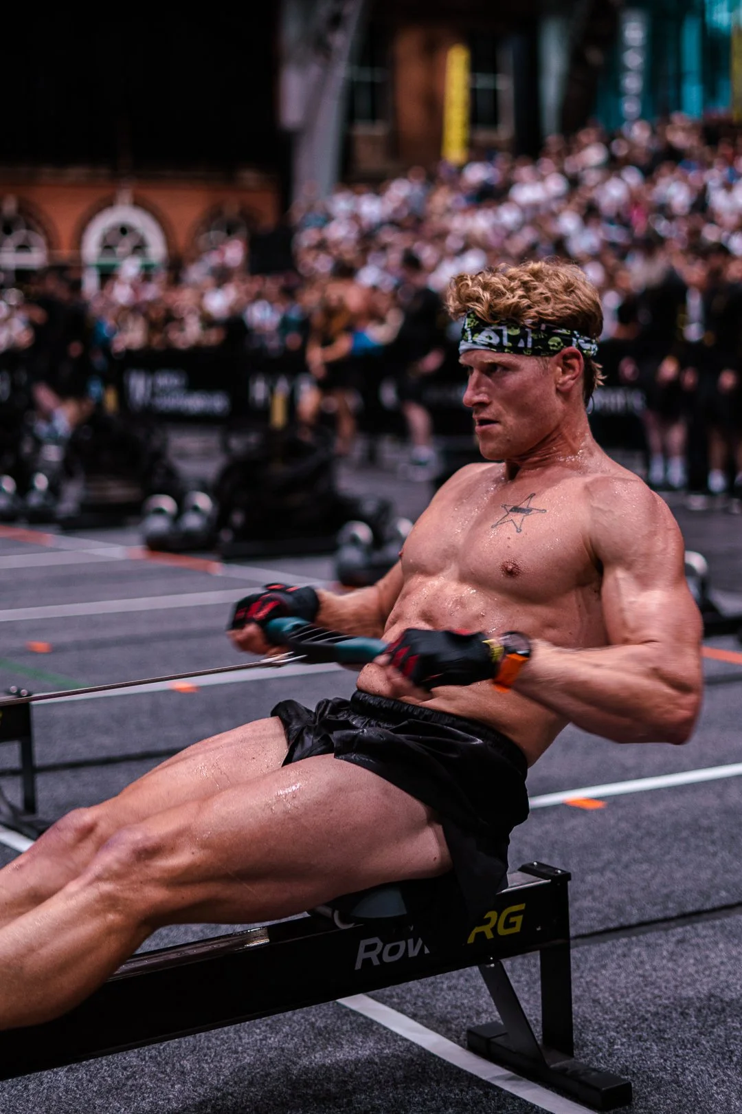 A muscular, shirtless man with a headband and black shorts exercises on a rowing machine in a gym, with a large crowd of people in the background.