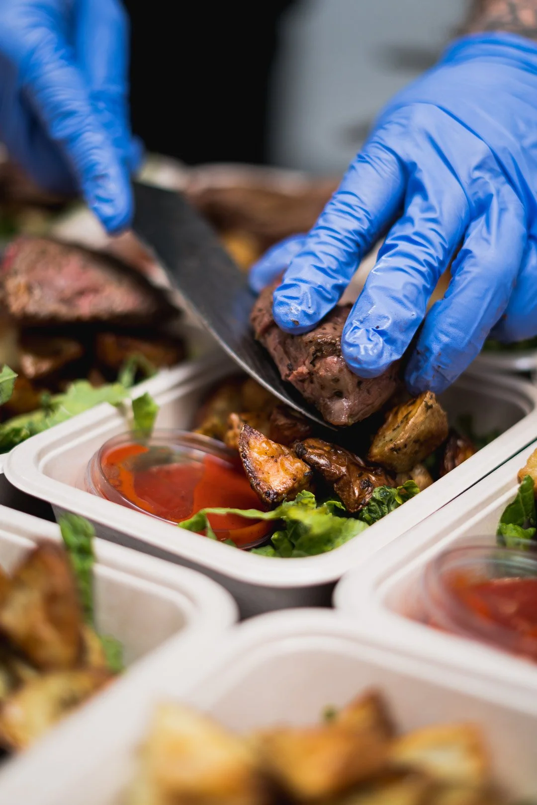 Person wearing blue gloves arranging grilled meat and vegetables in a takeaway container, with small bowls of sauce.