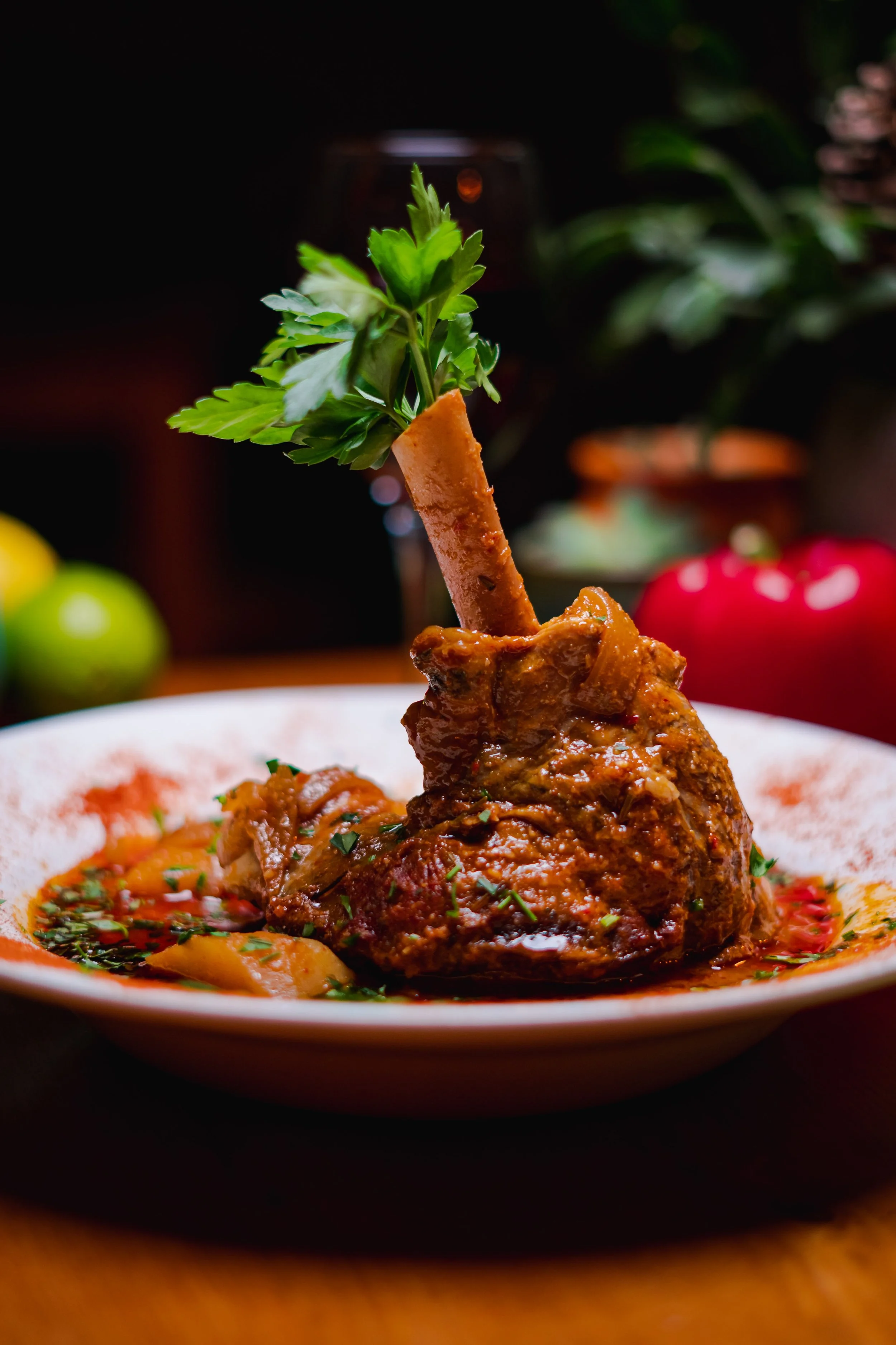 A cooked lamb shank garnished with parsley on a plate with sauce and vegetables, with apples and greens in the background.