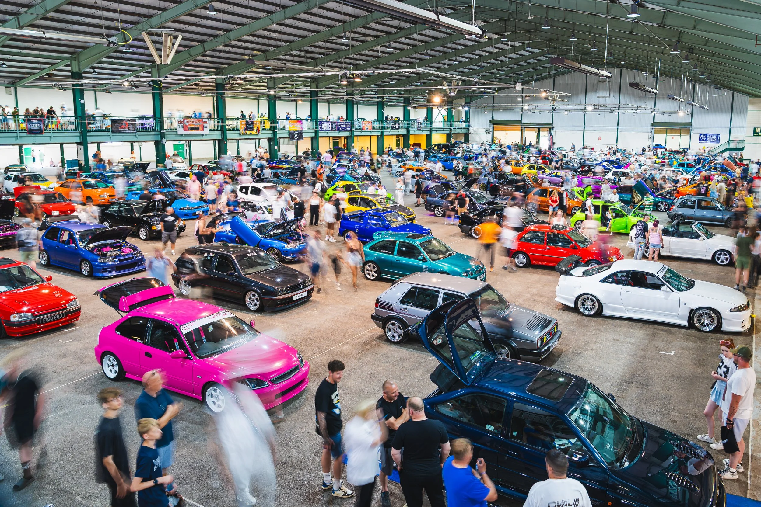 Indoor car exhibition showcasing a variety of colorful customized cars with many visitors walking around and viewing the vehicles.