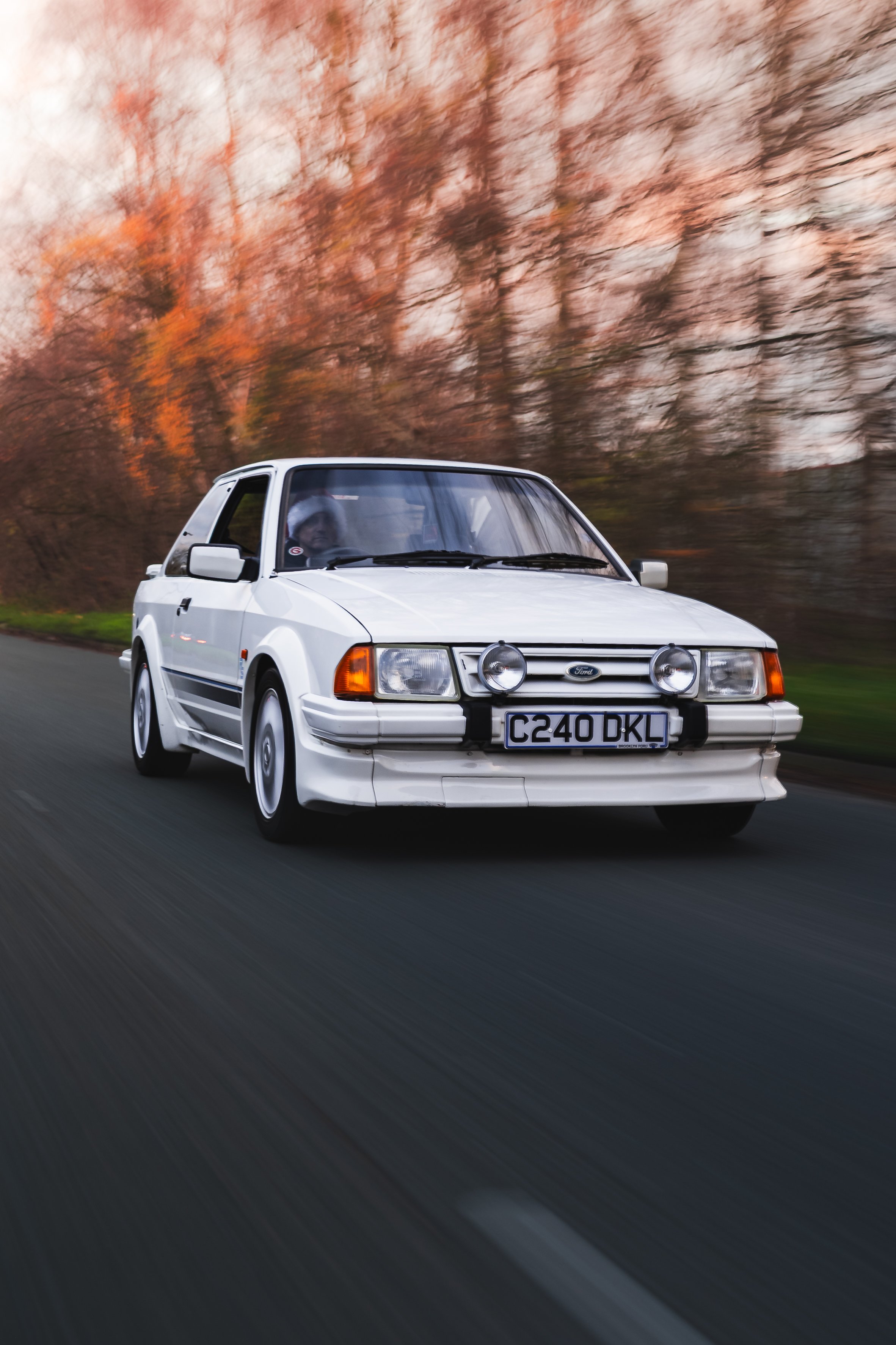 A white vintage Ford rally car driving on a road with autumn trees in the background.