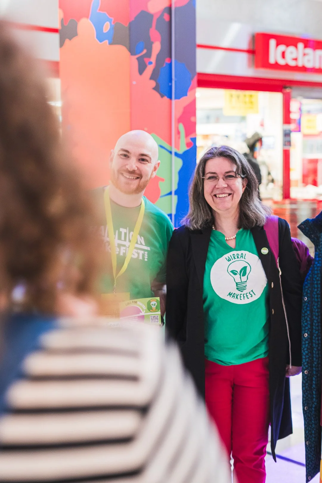 People smiling at the Wirral Makefest event, with a colorful background and a woman wearing the event logo on her shirt.