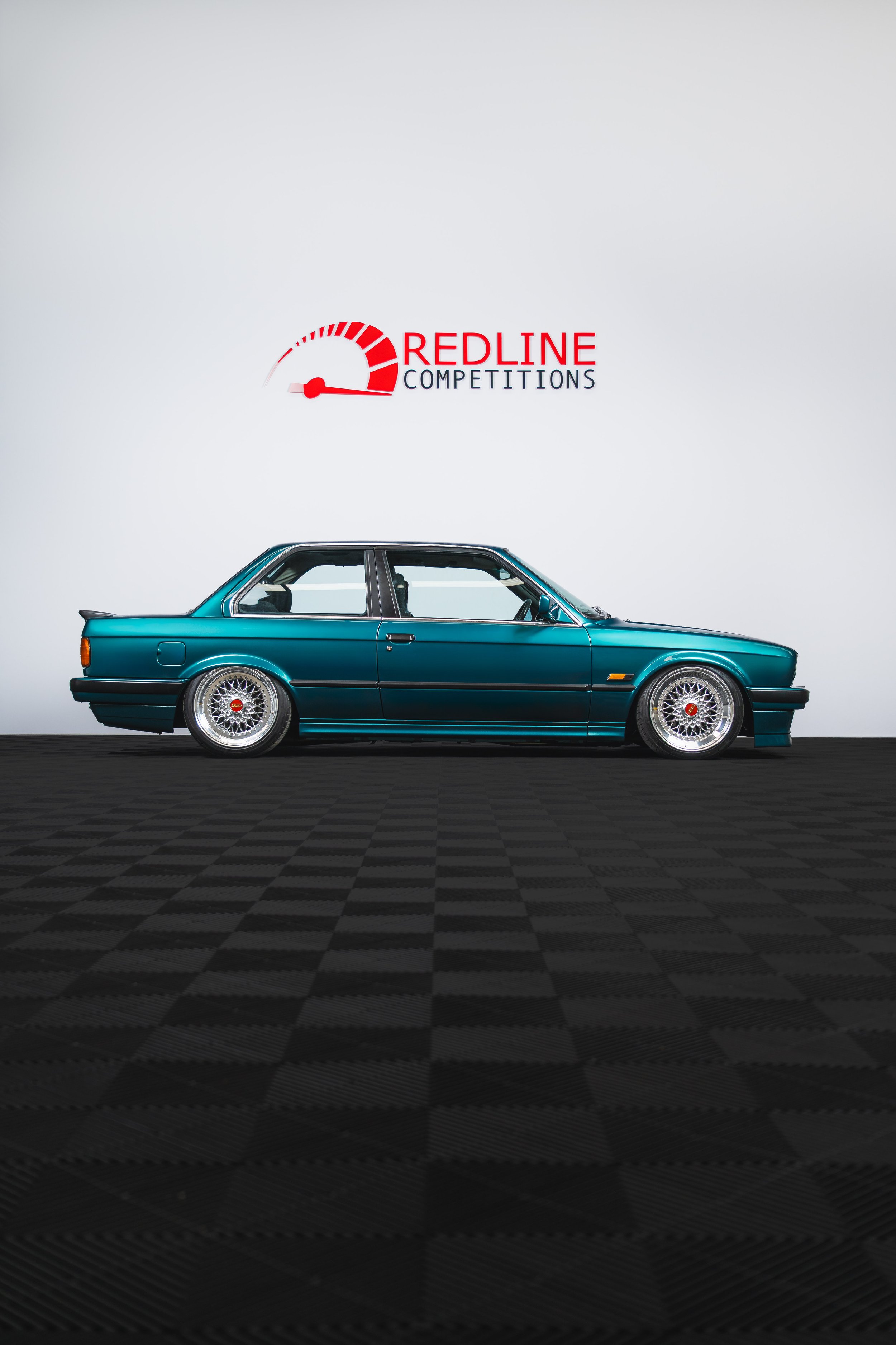 A teal vintage car with custom wheels is positioned sideways on a black checkered floor in front of a white wall displaying the Redline Competitions logo.