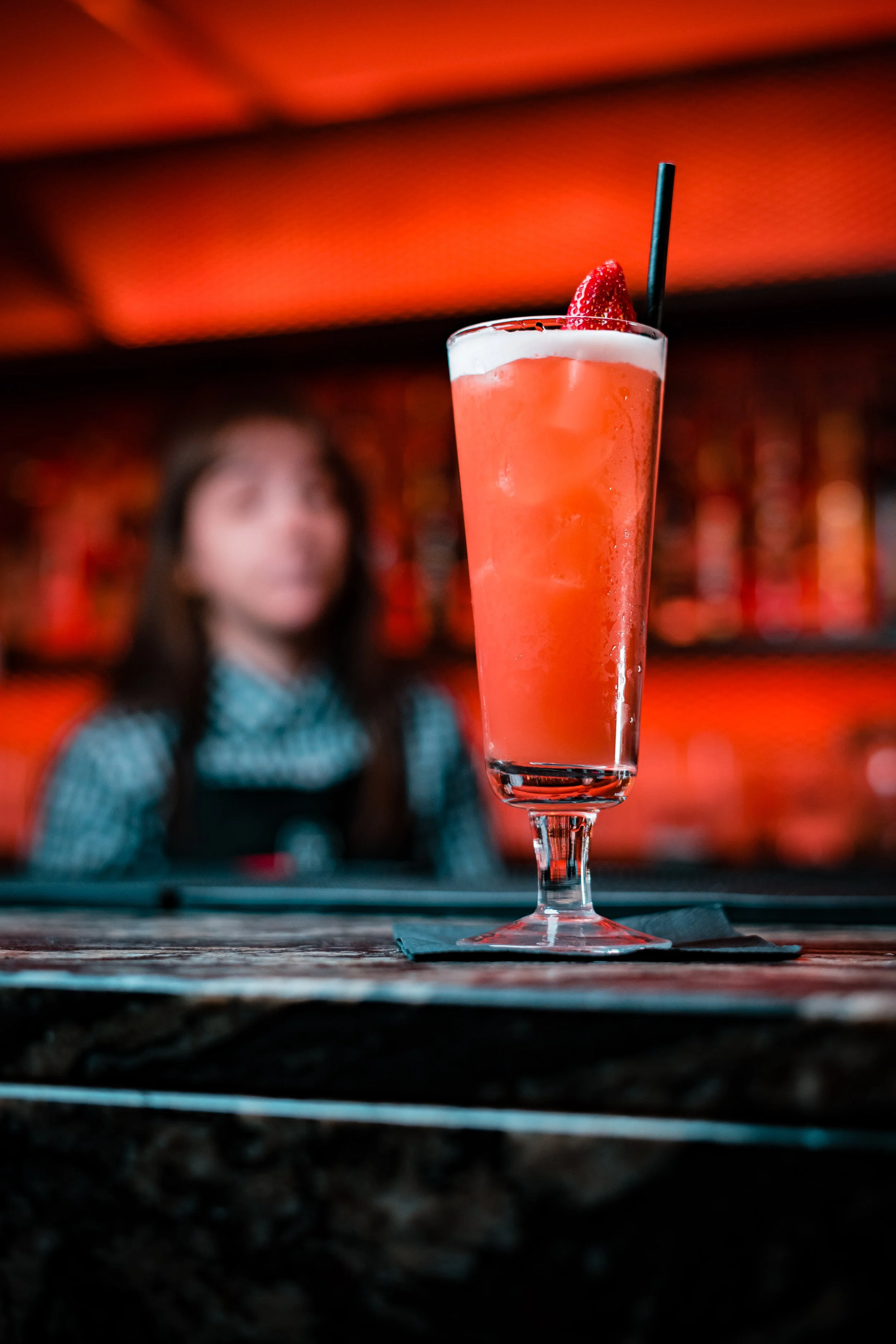 A pink cocktail garnished with a strawberry and a black straw on a bar counter, with a bartender in blurred background.