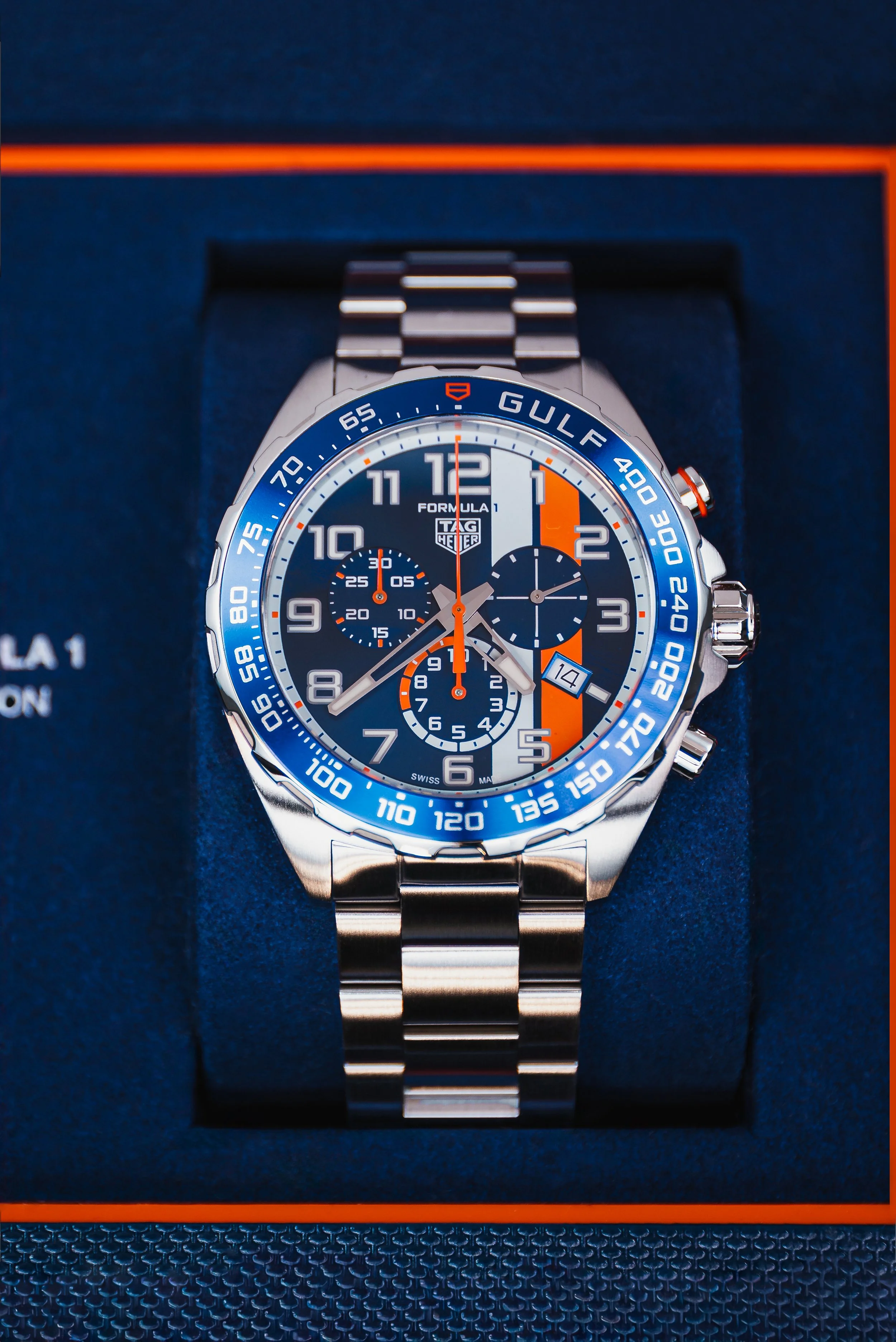 A TAG Heuer Formula 1 chronograph wristwatch with a stainless steel band, blue dial, multiple sub-dials, and a tachymeter bezel, displayed in a dark blue box with orange accents.
