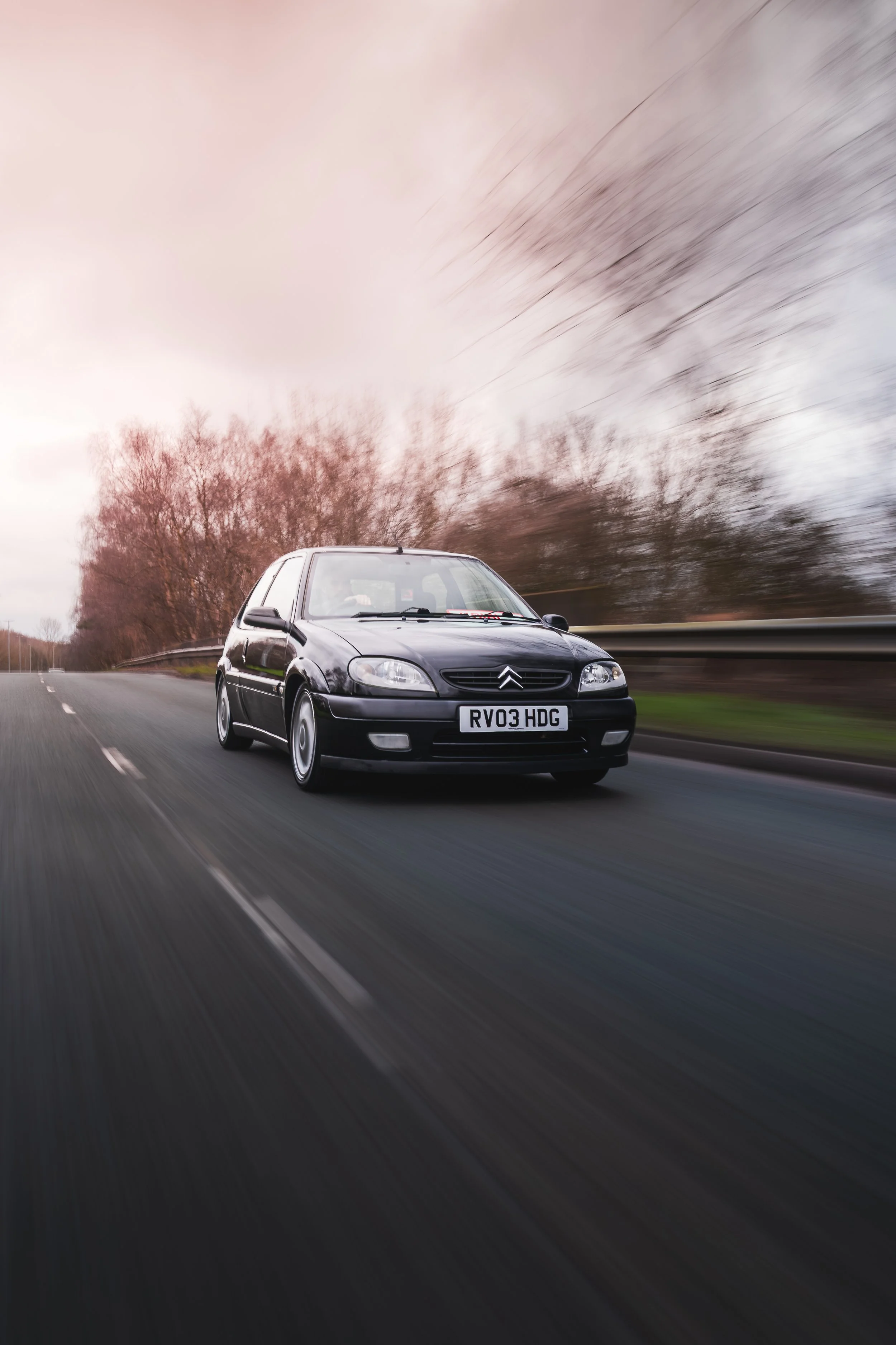 A black Citroën hatchback car driving on a highway with motion blur, overcast sky, and trees in the background.