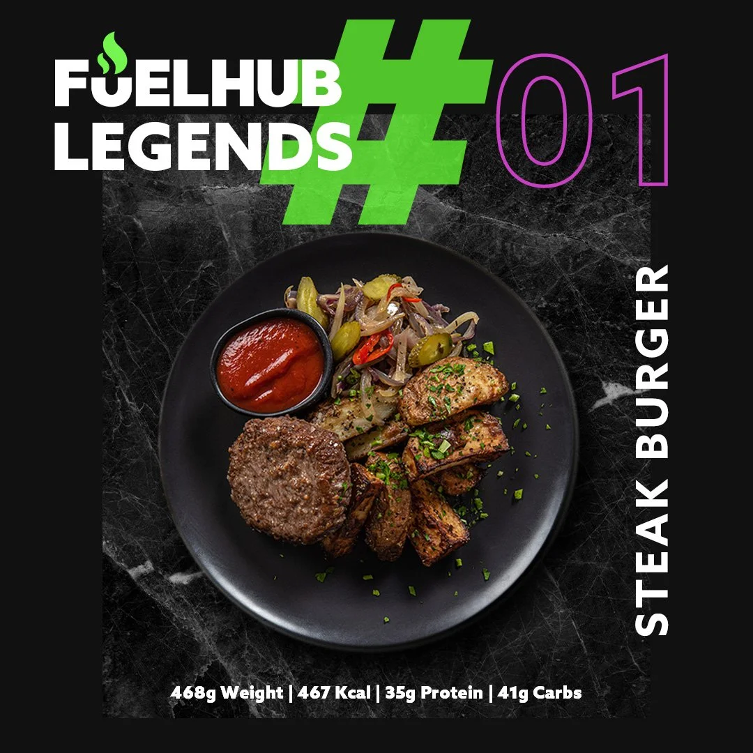 A black plate with a hamburger patty, cooked steak pieces, sautéed vegetables, and a small cup of ketchup on a dark surface. The image features overlaid text promoting Fuelhub Legends, with nutritional info listed below.