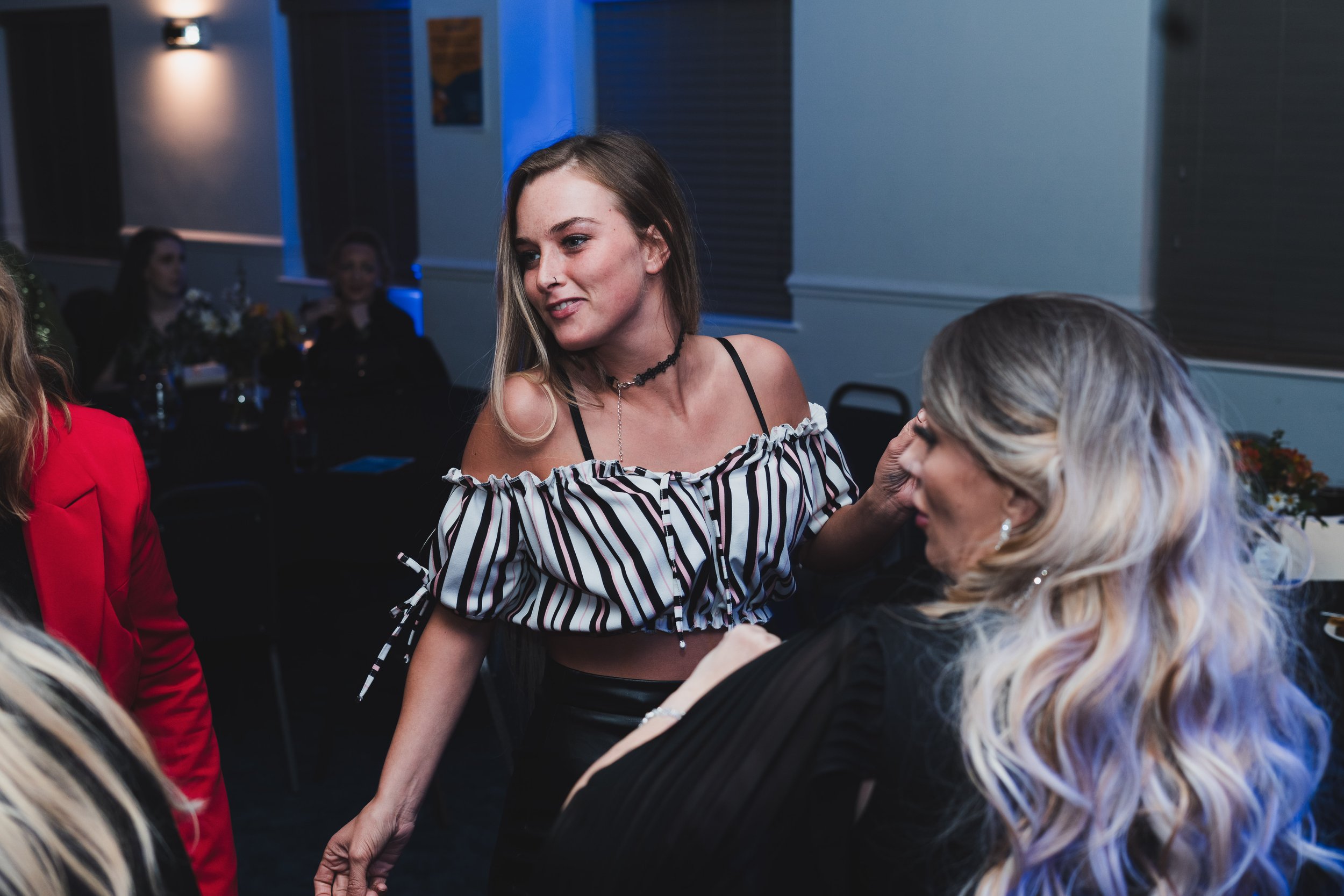 Young woman with long light brown hair, wearing a black choker and off-shoulder striped top, smiling while interacting with an older woman with white hair. They are at a social gathering in a dimly lit room with blue lighting and other people in the 
