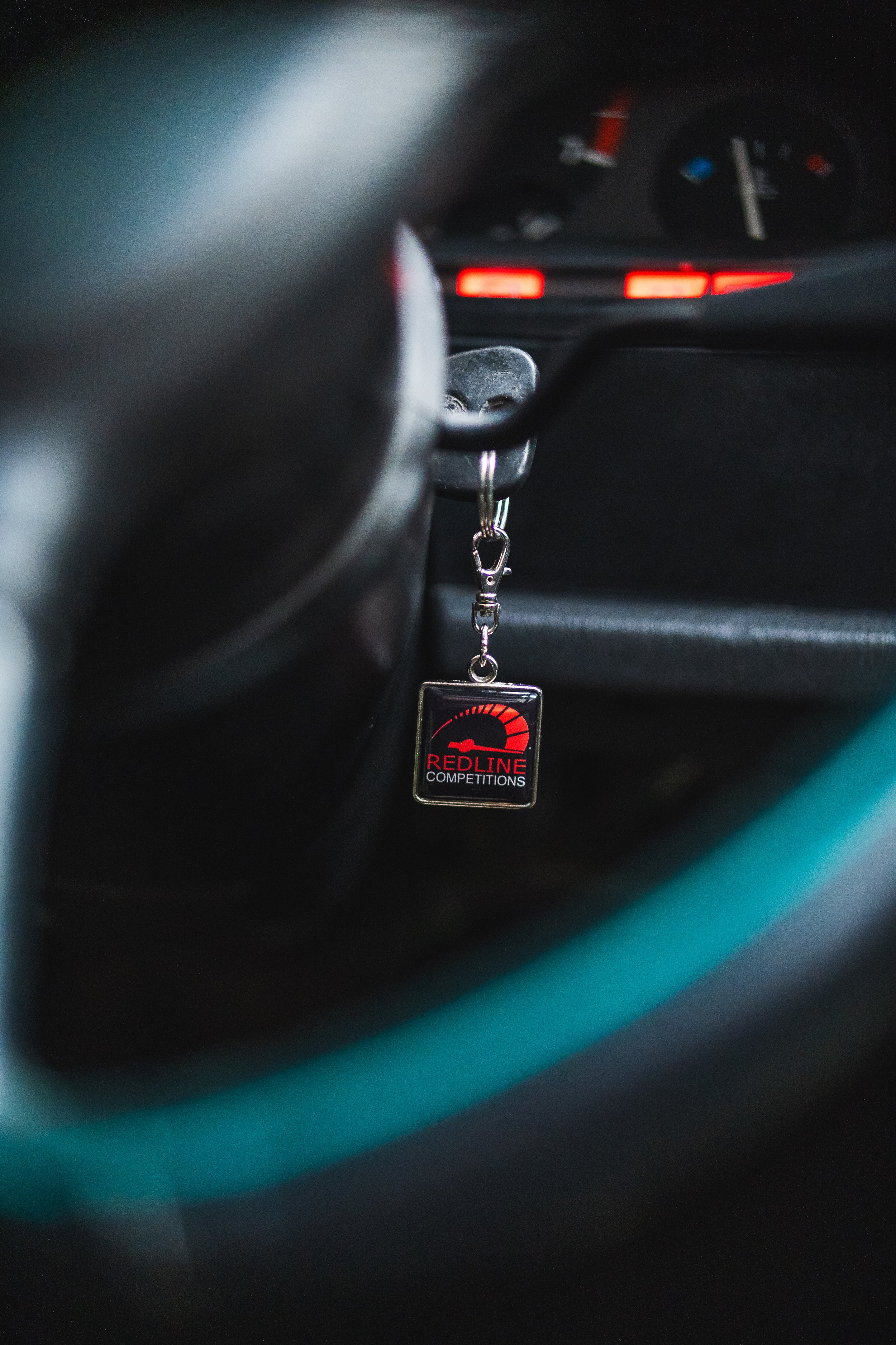Close-up of a car interior showing a keychain hanging from the ignition, with a logo that reads 'Redline Competitions' and a speedometer graphic.