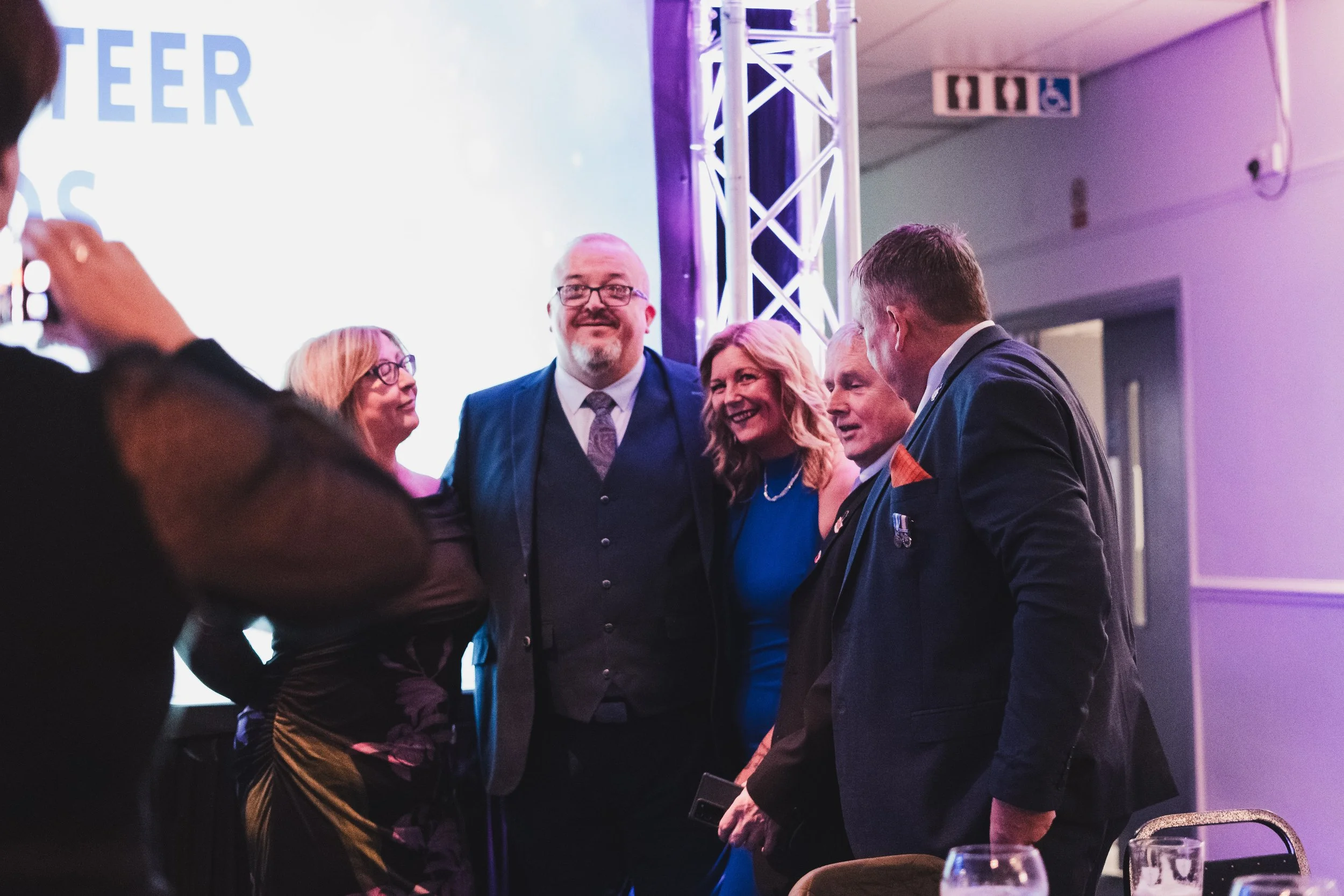 Group of five adults smiling and talking at a formal event, with a large screen and structural lighting in the background.