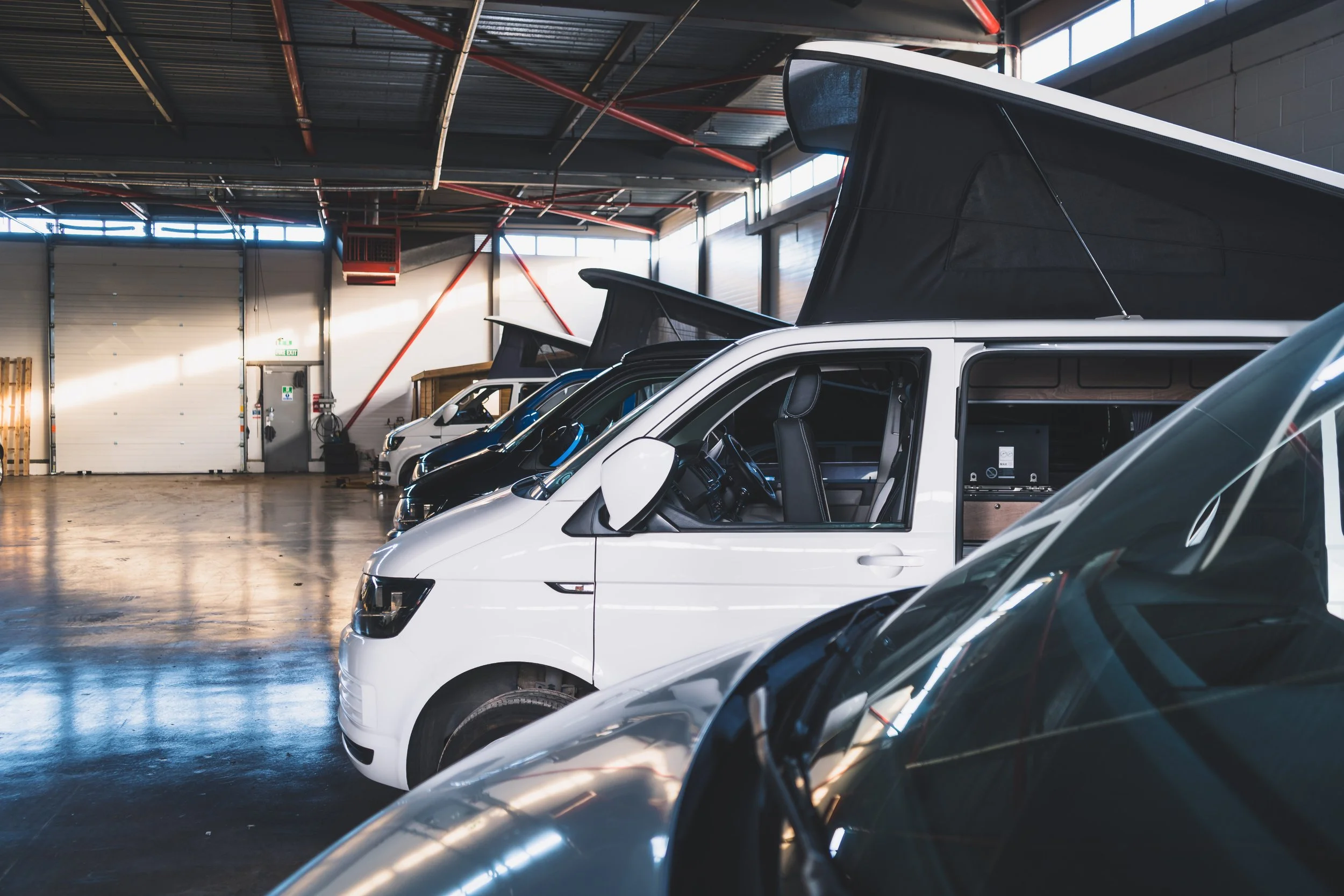 Multiple electric vehicles with black and white camper van roofs parked inside a spacious warehouse with high ceilings and large garage doors.