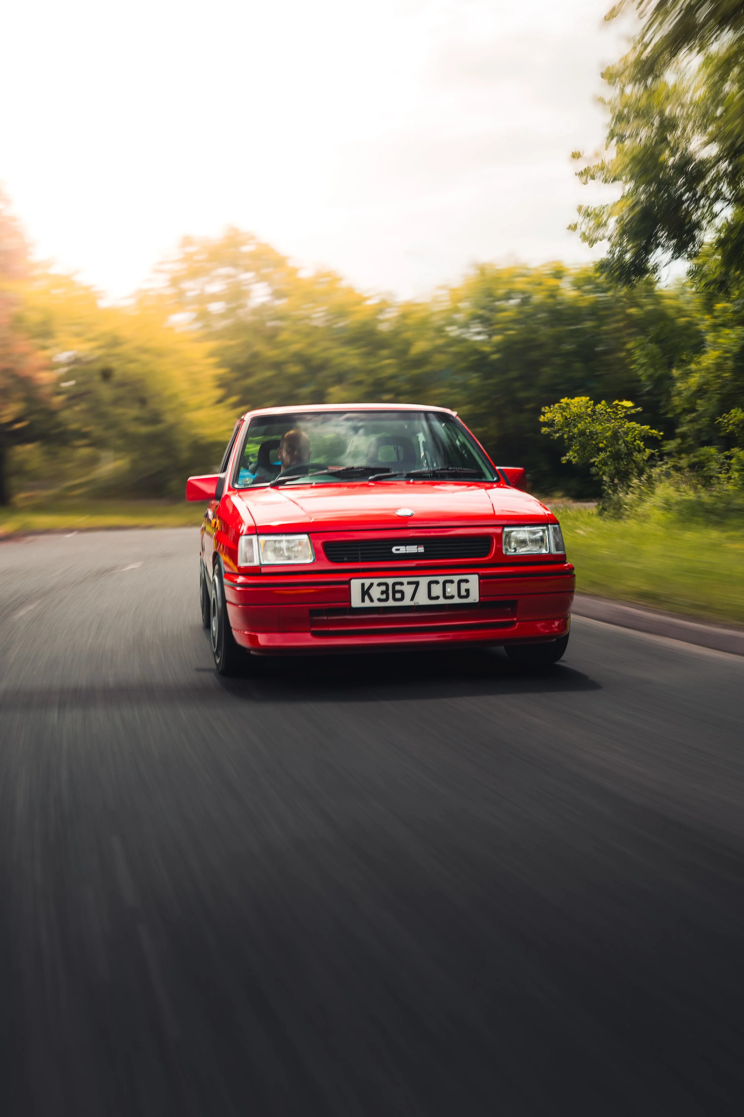 A red vintage car driving on a winding road surrounded by green trees, with motion blur indicating speed.