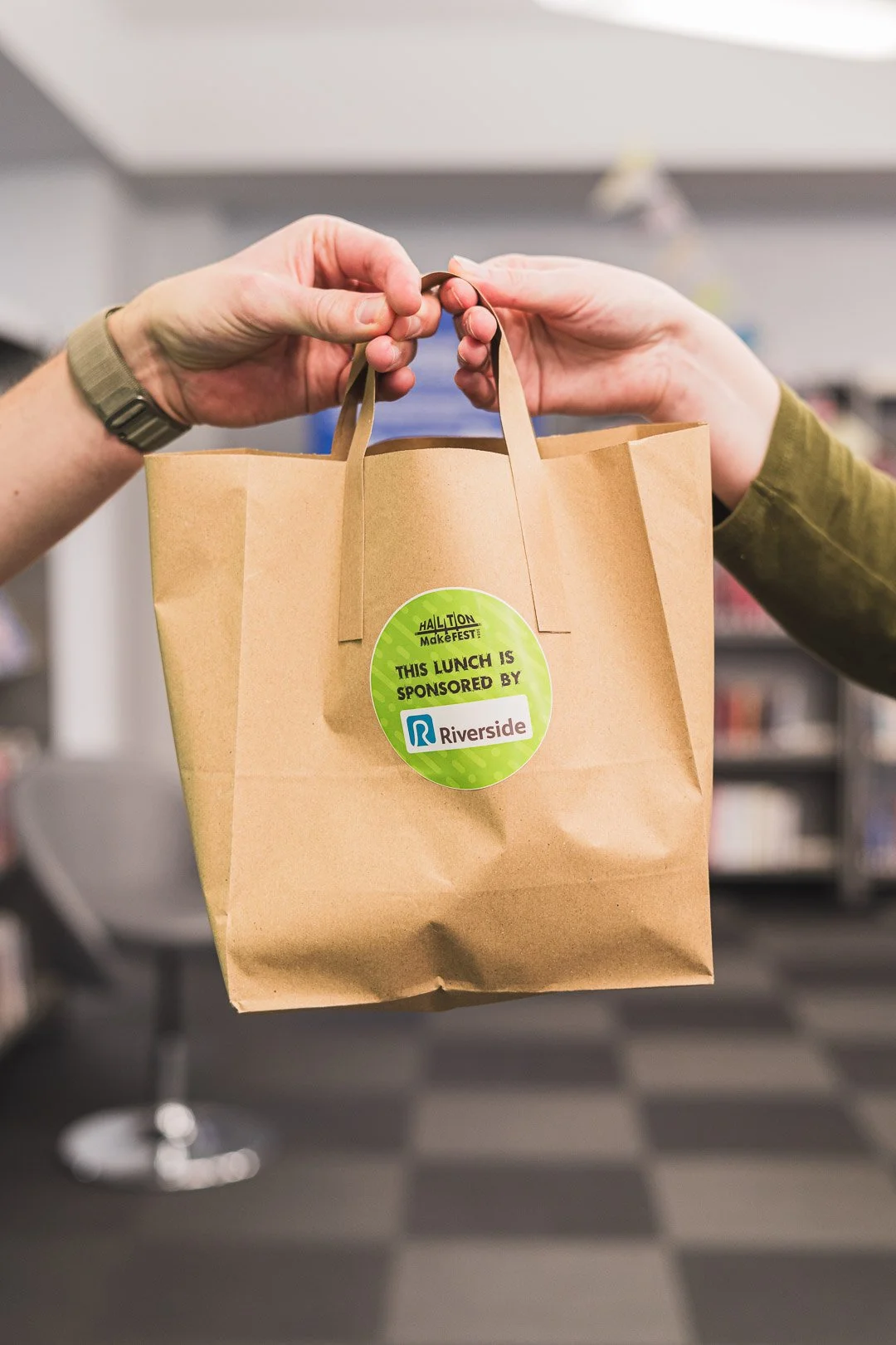 Two hands holding a brown paper lunch bag with a green circular sticker that reads, "This lunch is sponsored by Riverside," in an indoor setting.