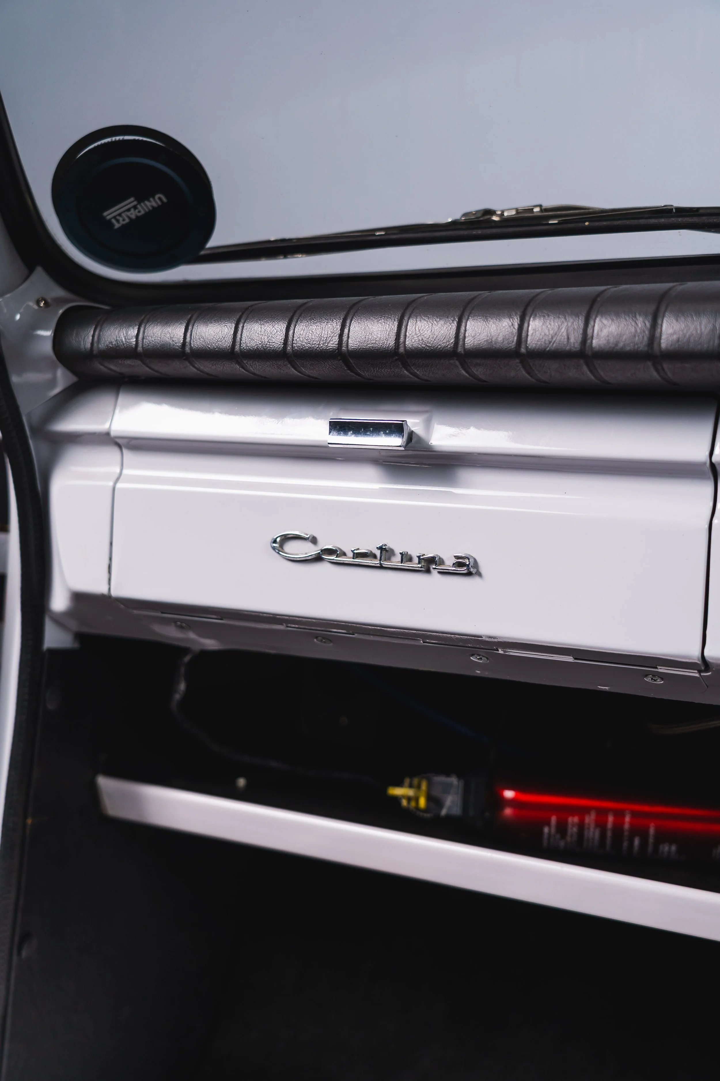 The dashboard of a vintage Toyota Corona with a black leather padded trim, a mirror with the Toyota logo, and a white glove box labeled 'Corona'.