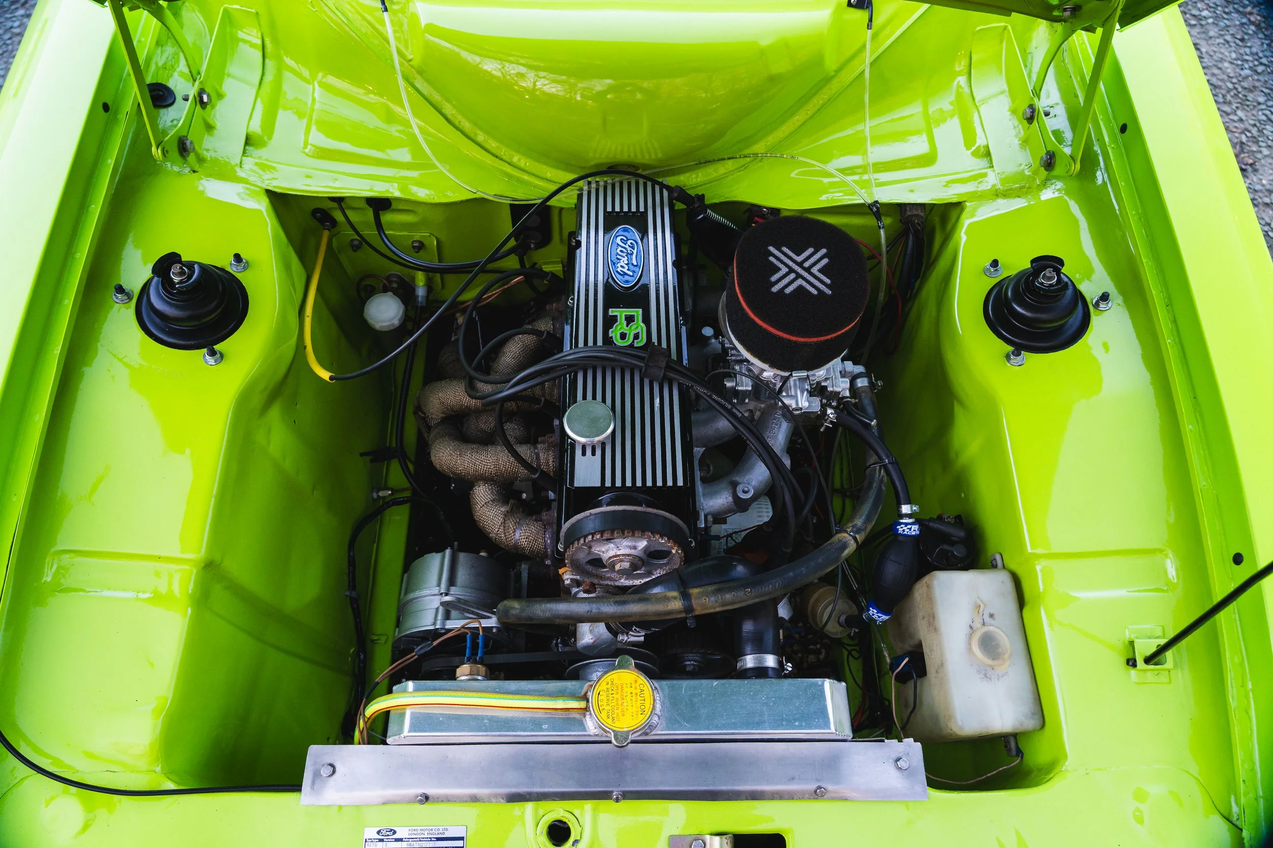 View of a car engine in a bright green engine bay, featuring a black Ford RS engine, an air filter, various hoses, wires, and mechanical components.