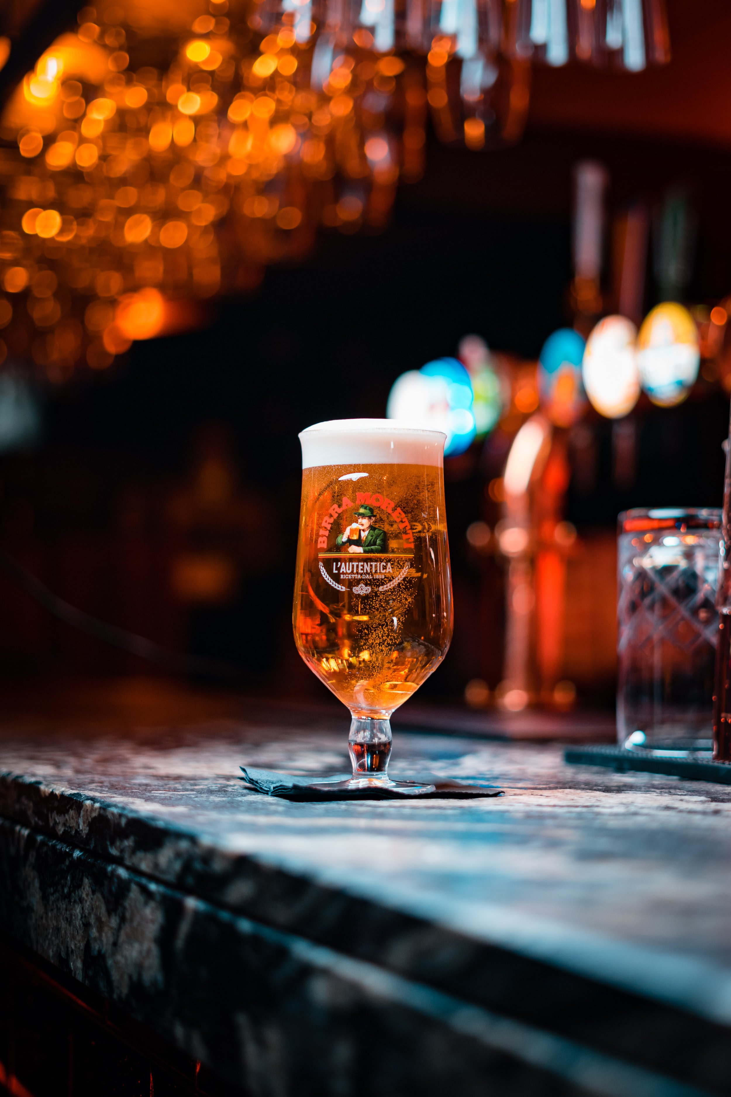 A glass of beer with foam on top sitting on a wooden bar counter with blurred colorful lights in the background.