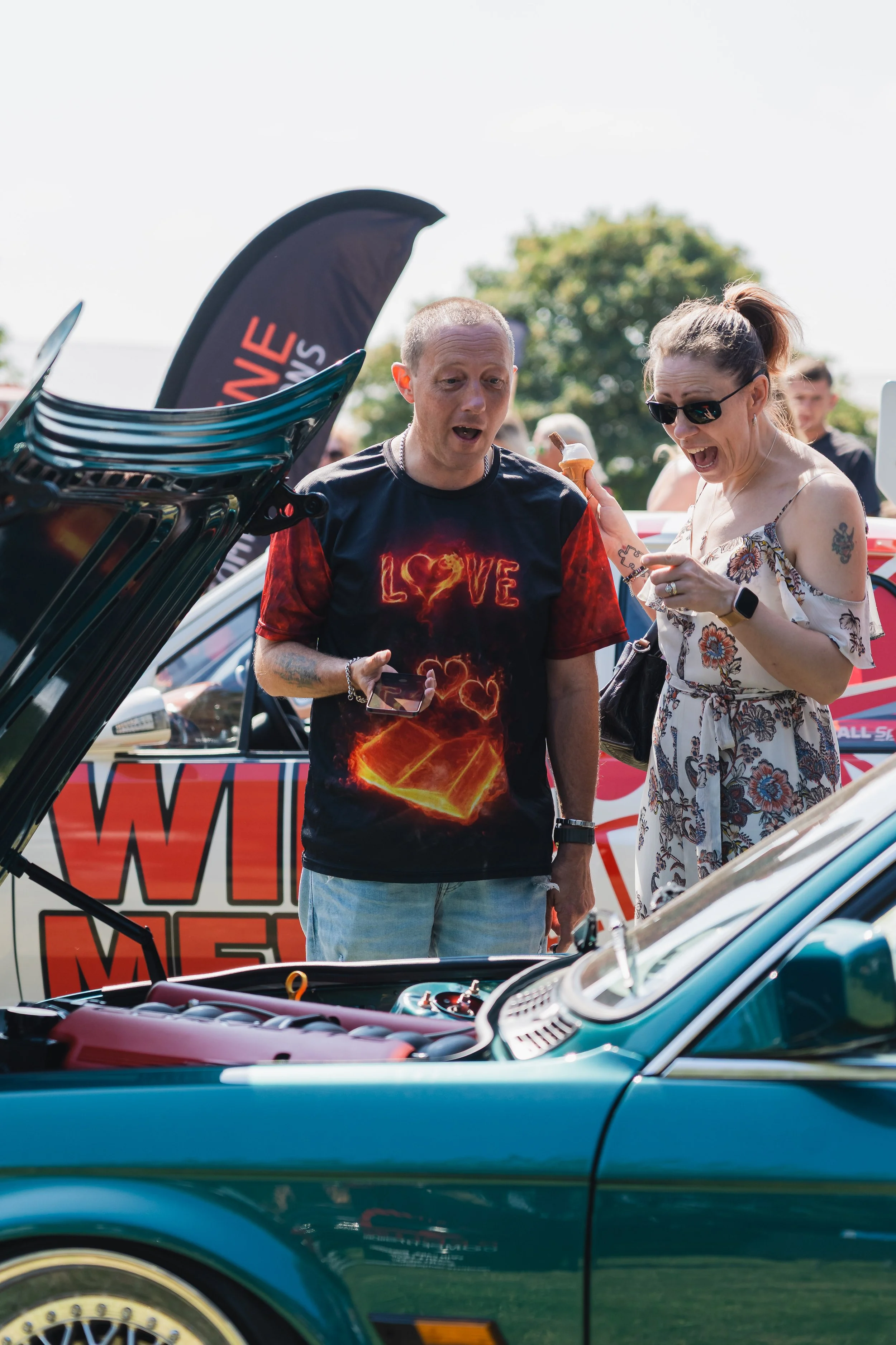 Two people are at a car show, looking at the engine of a teal classic car. The man has short hair and is wearing a black T-shirt with a fiery love design, holding a phone. The woman has glasses, tied-up hair, a tattoo on her arm, and is wearing a flo