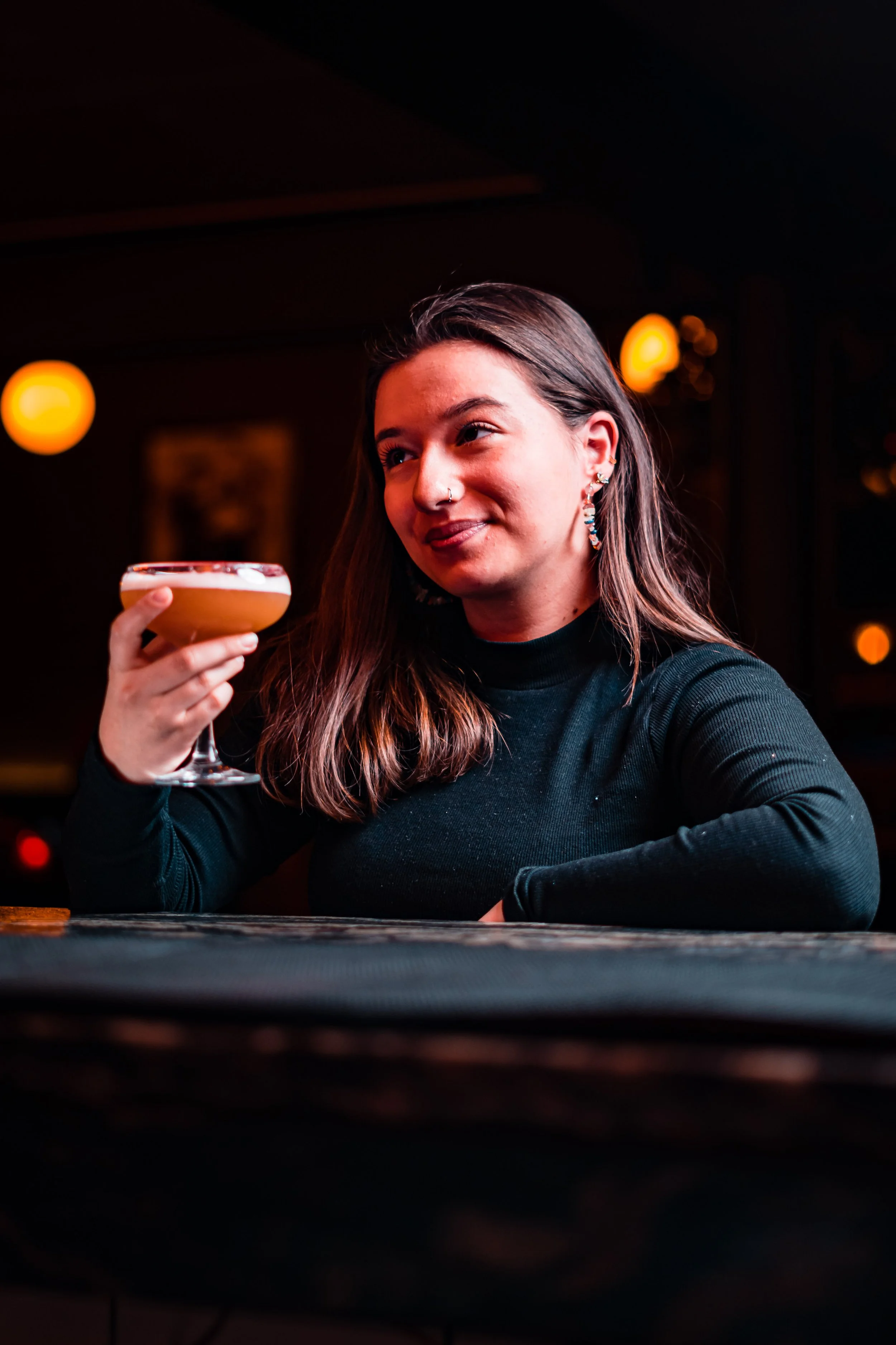 A young woman in a black top sitting at a bar holding a cocktail in a coupe glass, smiling and looking slightly to the side in a dimly-lit bar or restaurant.