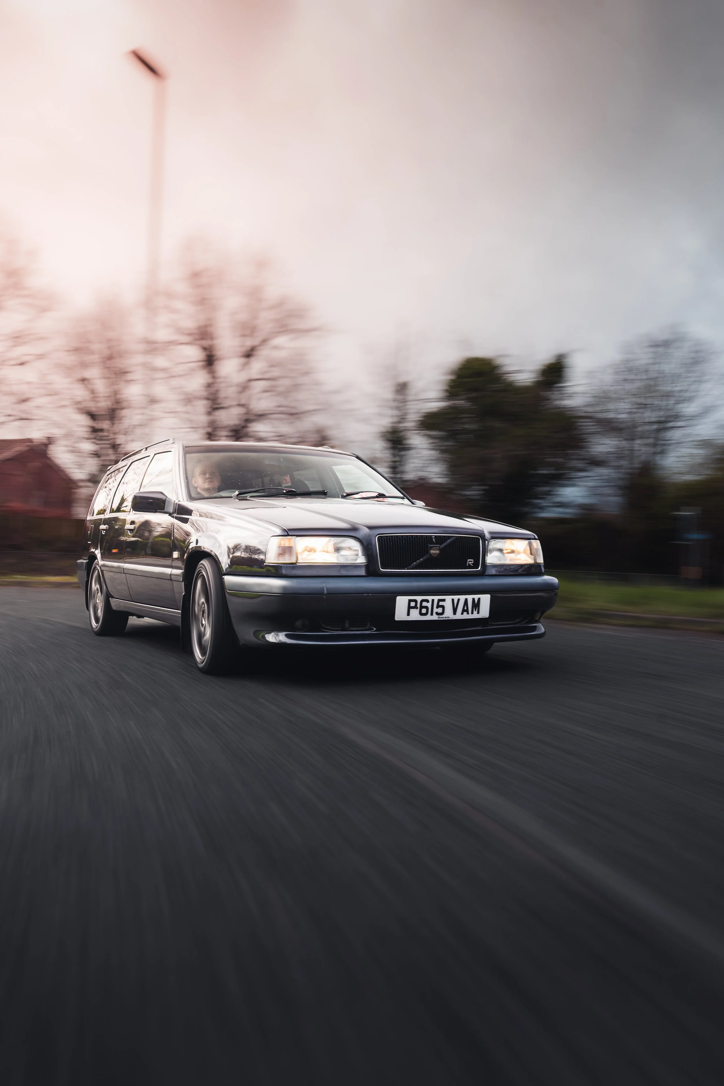 A black vintage Volvo station wagon driving on a road with a blurred background of trees and houses, during dusk or dawn.