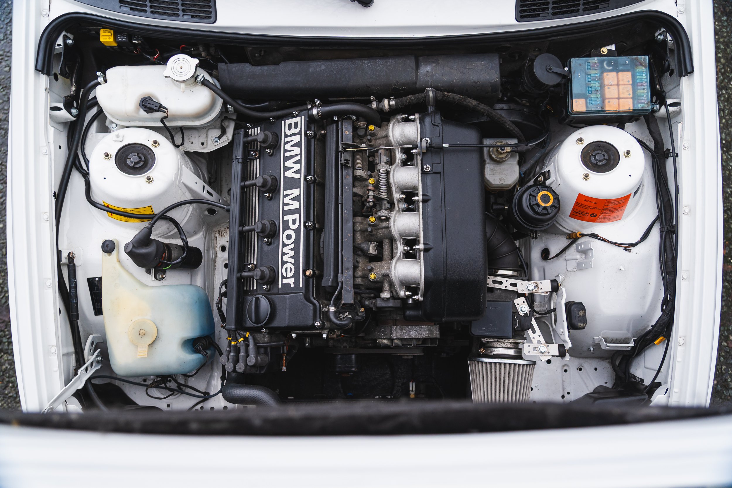 Top view of a BMW car engine with various components, hoses, and fluid reservoirs visible.