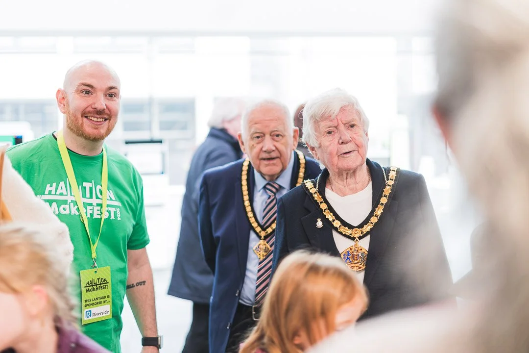 Group of people, including an elderly woman wearing a chain of office, at an indoor event, some in formal attire and others in company shirts.