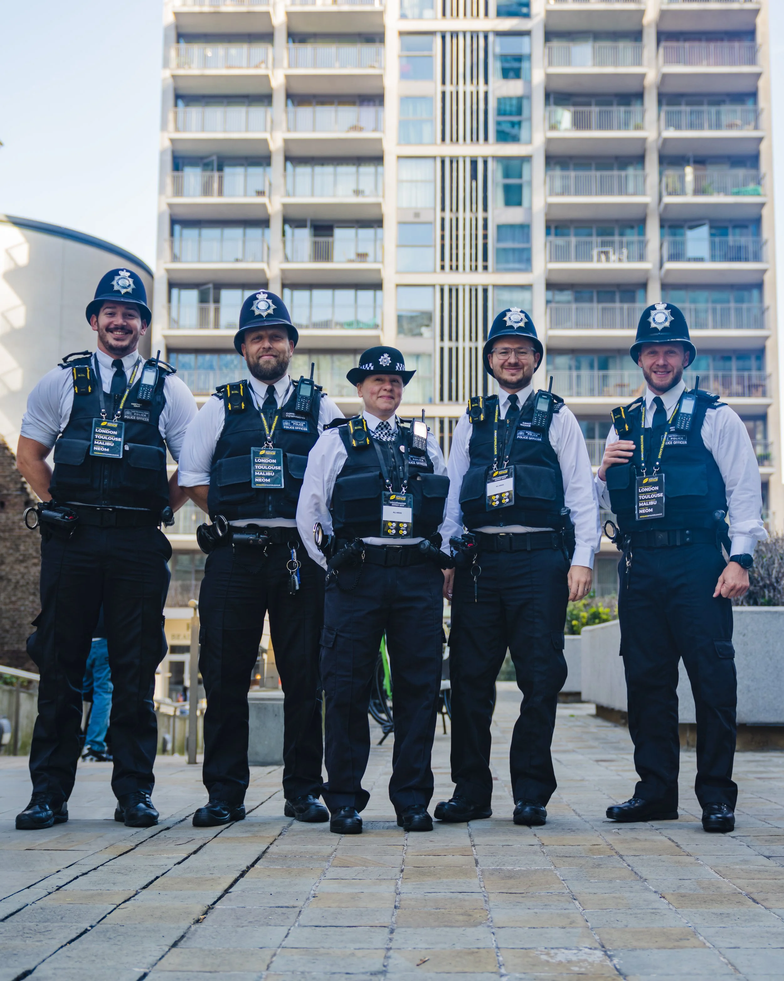 Five police officers standing in front of a high-rise building, wearing uniforms, hats, badges, and equipment, smiling at the camera.